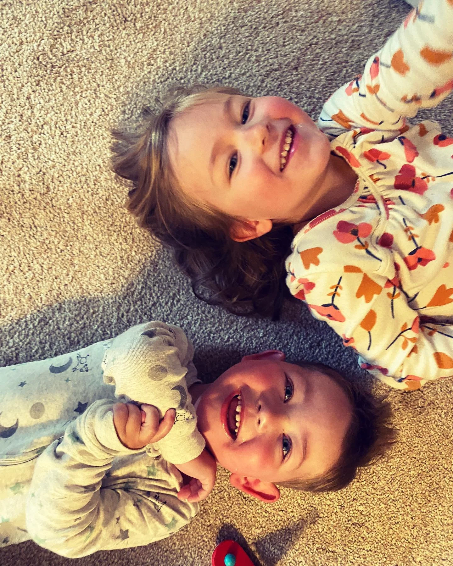 Two young children lying on a beige carpet, smiling and looking at the camera, one with curly hair and the other with straight hair, wearing pajamas with colorful patterns.