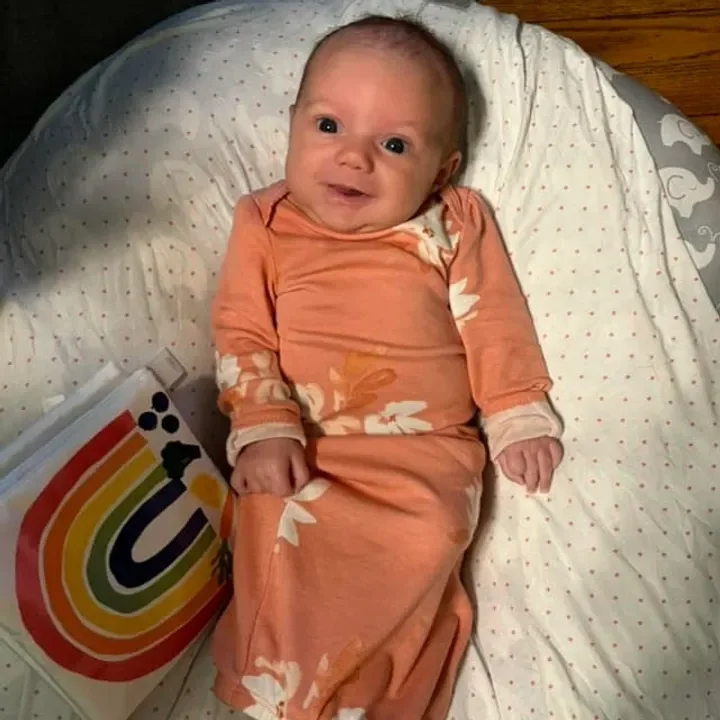 A smiling baby is lying on a white polka dot bedspread, wearing an orange dress with white floral patterns, next to a rainbow-themed book.