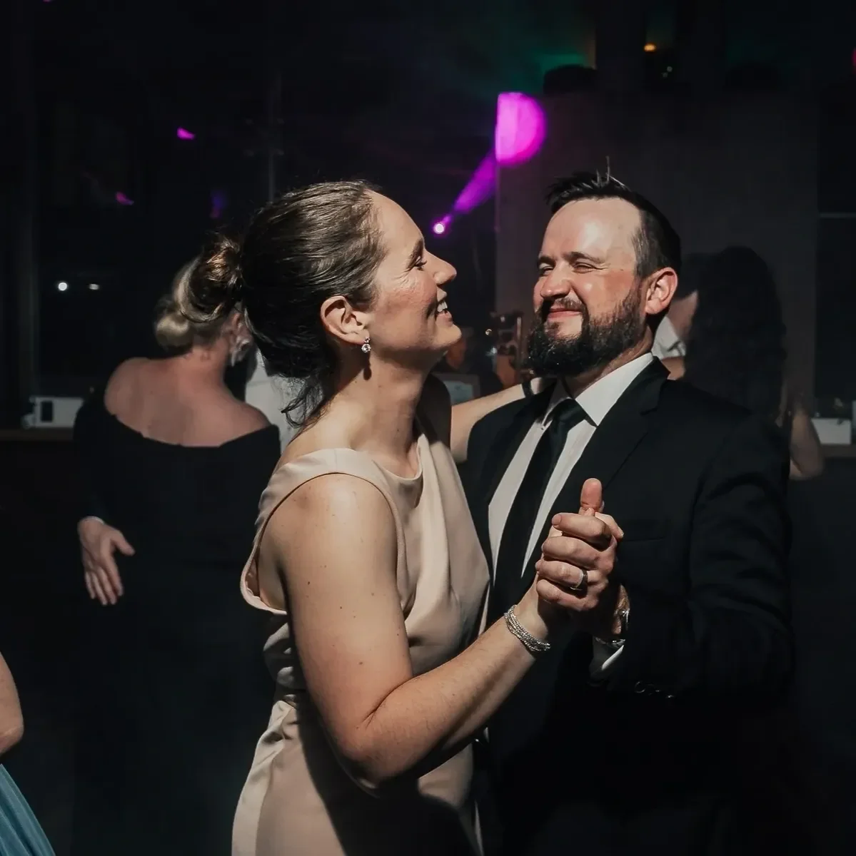 A smiling couple dancing at a formal event, with the woman wearing a beige dress and the man in a black suit and tie, in a dimly lit venue with pink and purple lighting.