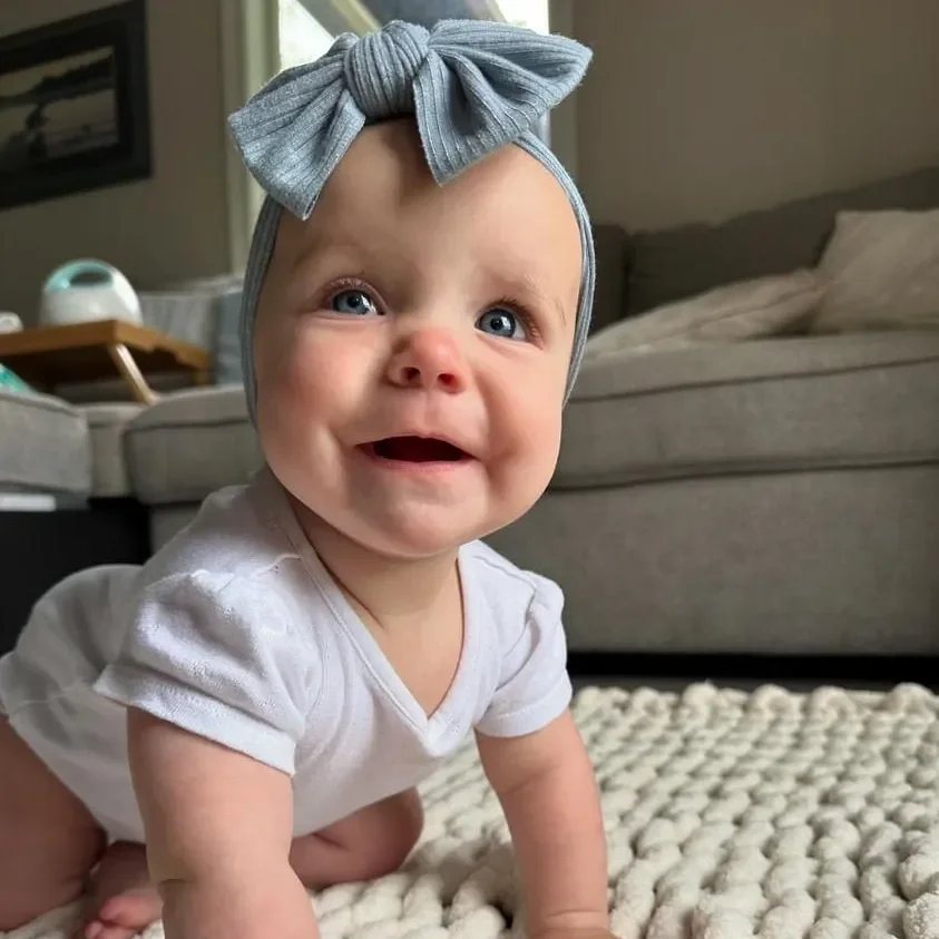 A smiling baby girl with blue eyes and a headband with a bow, crawling on a cozy textured rug in a living room.