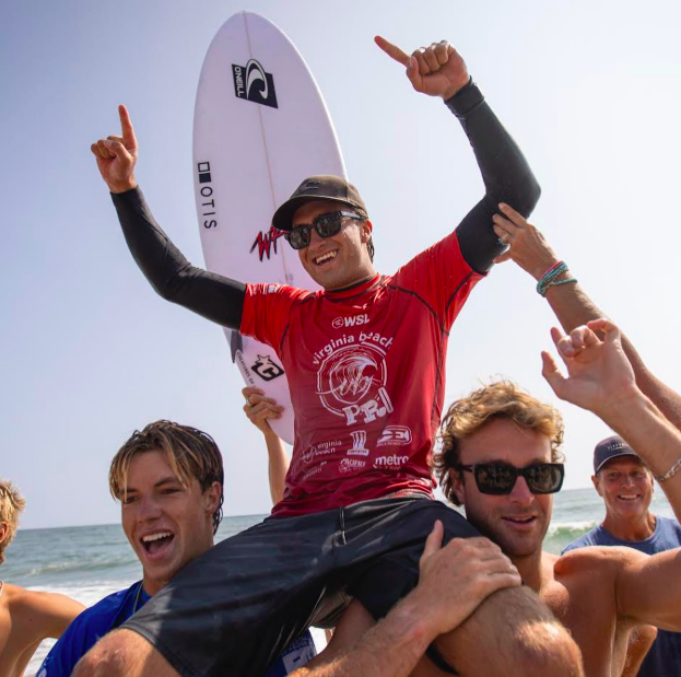 A man in a red T-shirt and sunglasses sitting on another person's shoulders at the beach, celebrating with his arms raised, holding a surfboard behind him, and surrounded by smiling friends.
