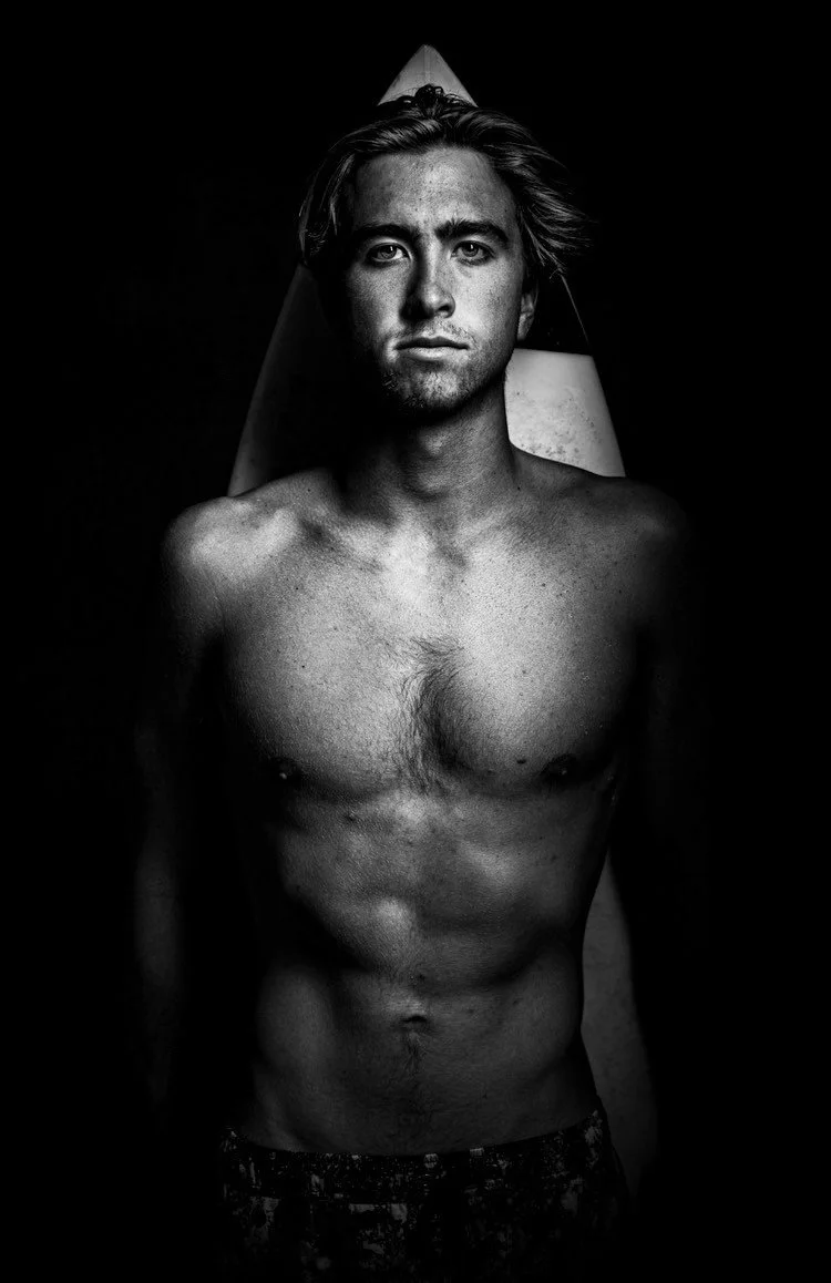 Black and white portrait of a shirtless young man with a serious expression, standing in front of a backdrop resembling a surfboard.