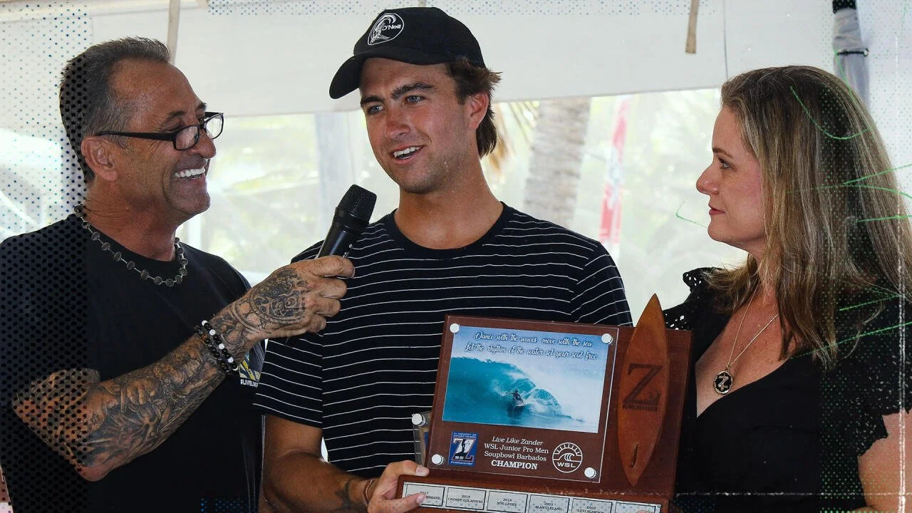 A man in a striped shirt holding an award plaque while being interviewed by an older man with tattoos and glasses, and a woman in black, in an outdoor setting