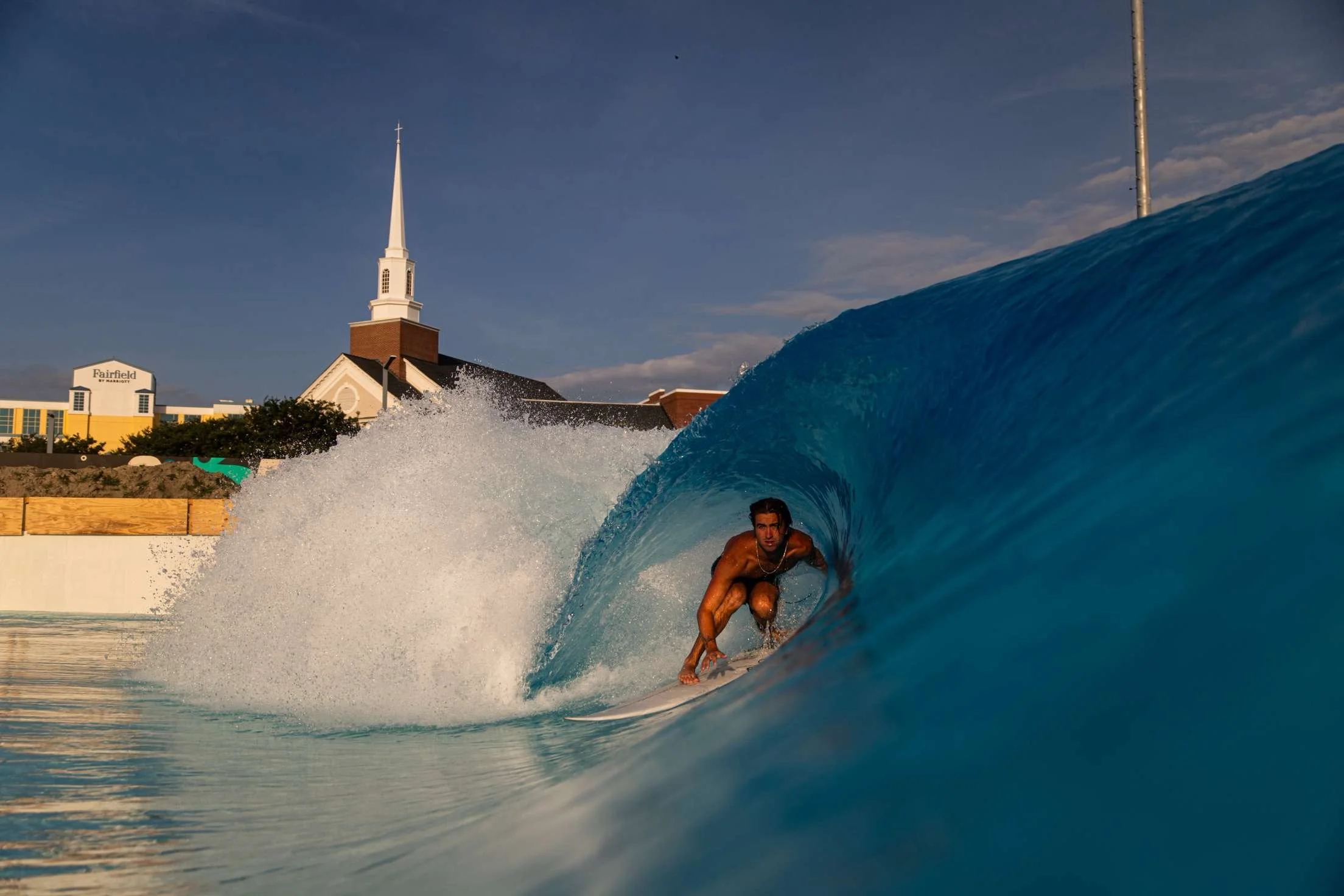 Male surfer riding inside the barrel of a breaking wave with buildings and a church steeple in the background.