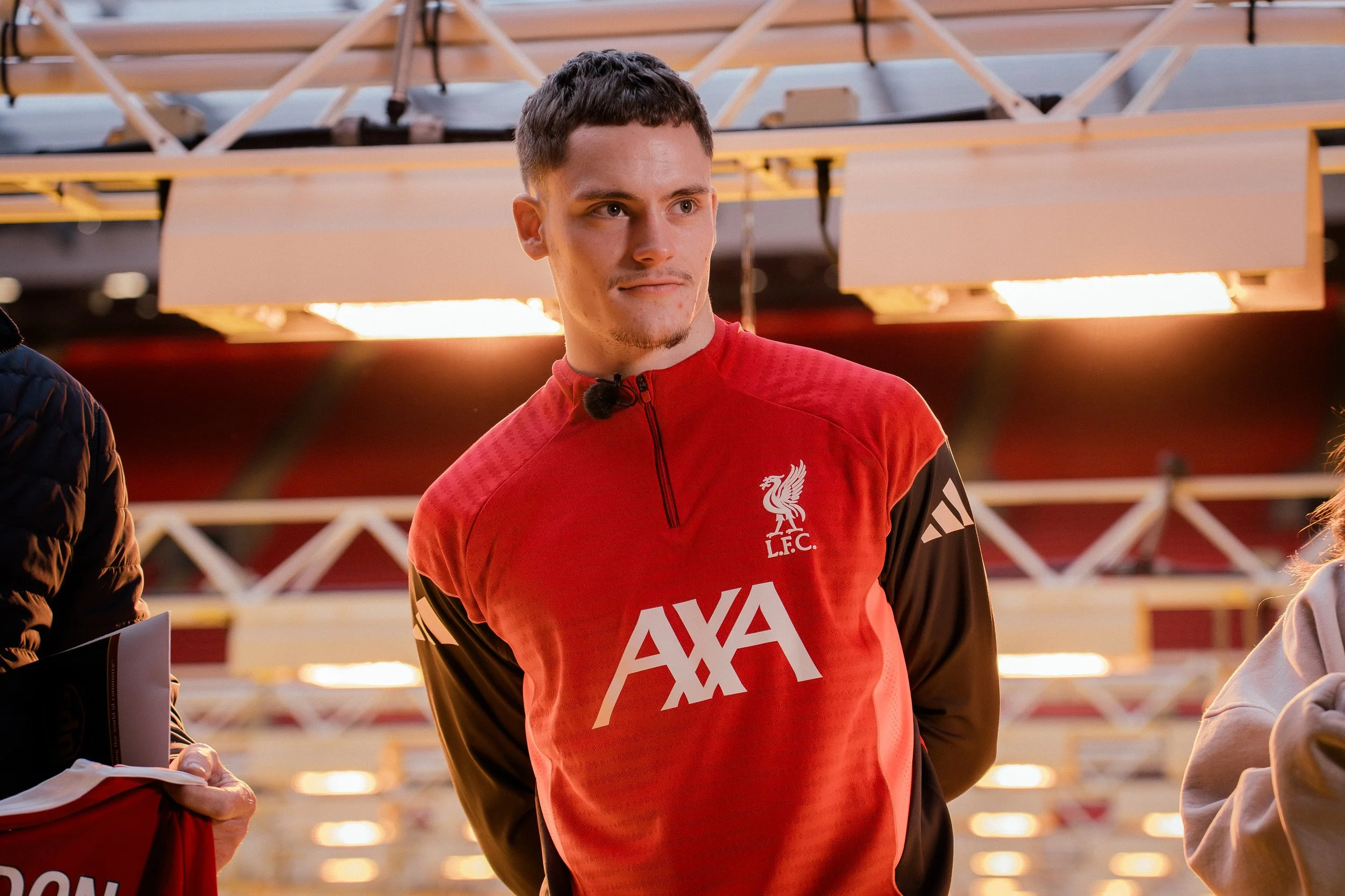 Florian Wirtz in a Liverpool Football Club soccer jersey stands with his hands behind his back, looking to his left, with an indoor stadium background.