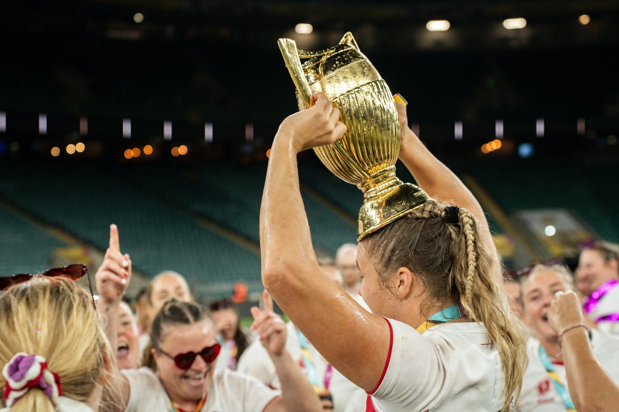 England Womens Rugby lifting a gold trophy above her head in celebration, surrounded by teammates in a stadium.