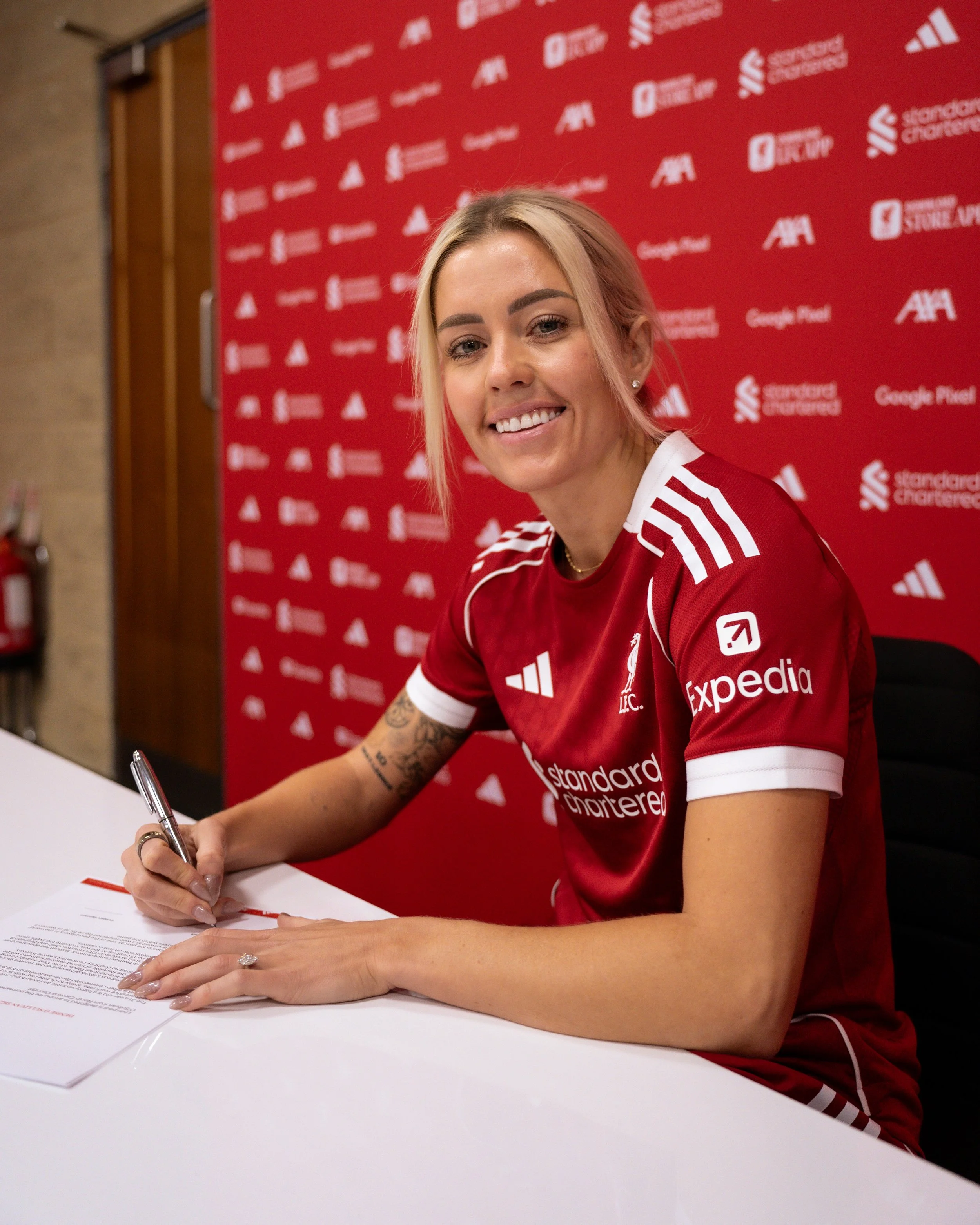 Denise O'Sullivan in a red soccer jersey is sitting at a table, smiling and signing papers.