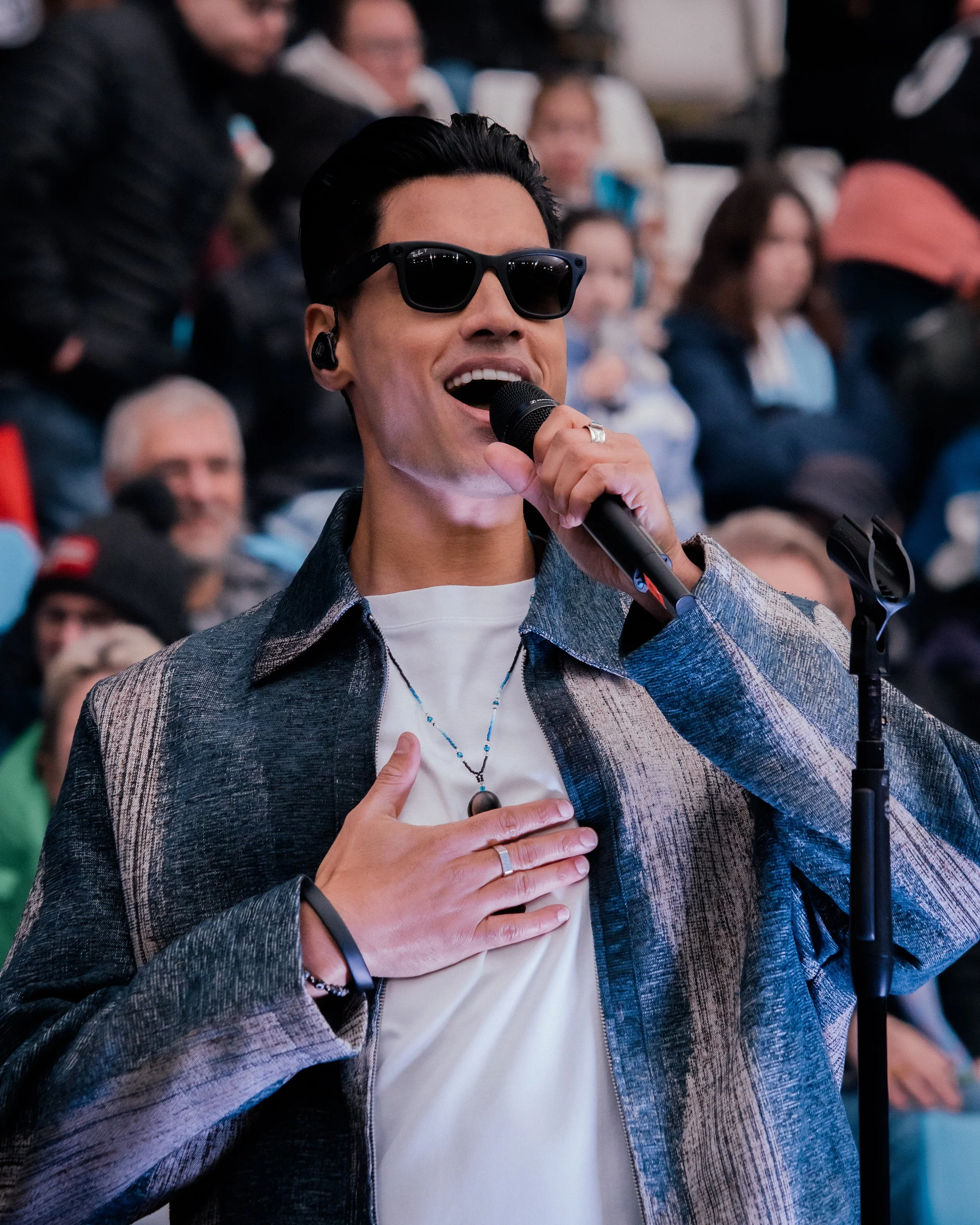 Siva Kaneswaran into a microphone with his hand over his heart, wearing sunglasses, a patterned jacket, and a white shirt, with a crowd in the background.