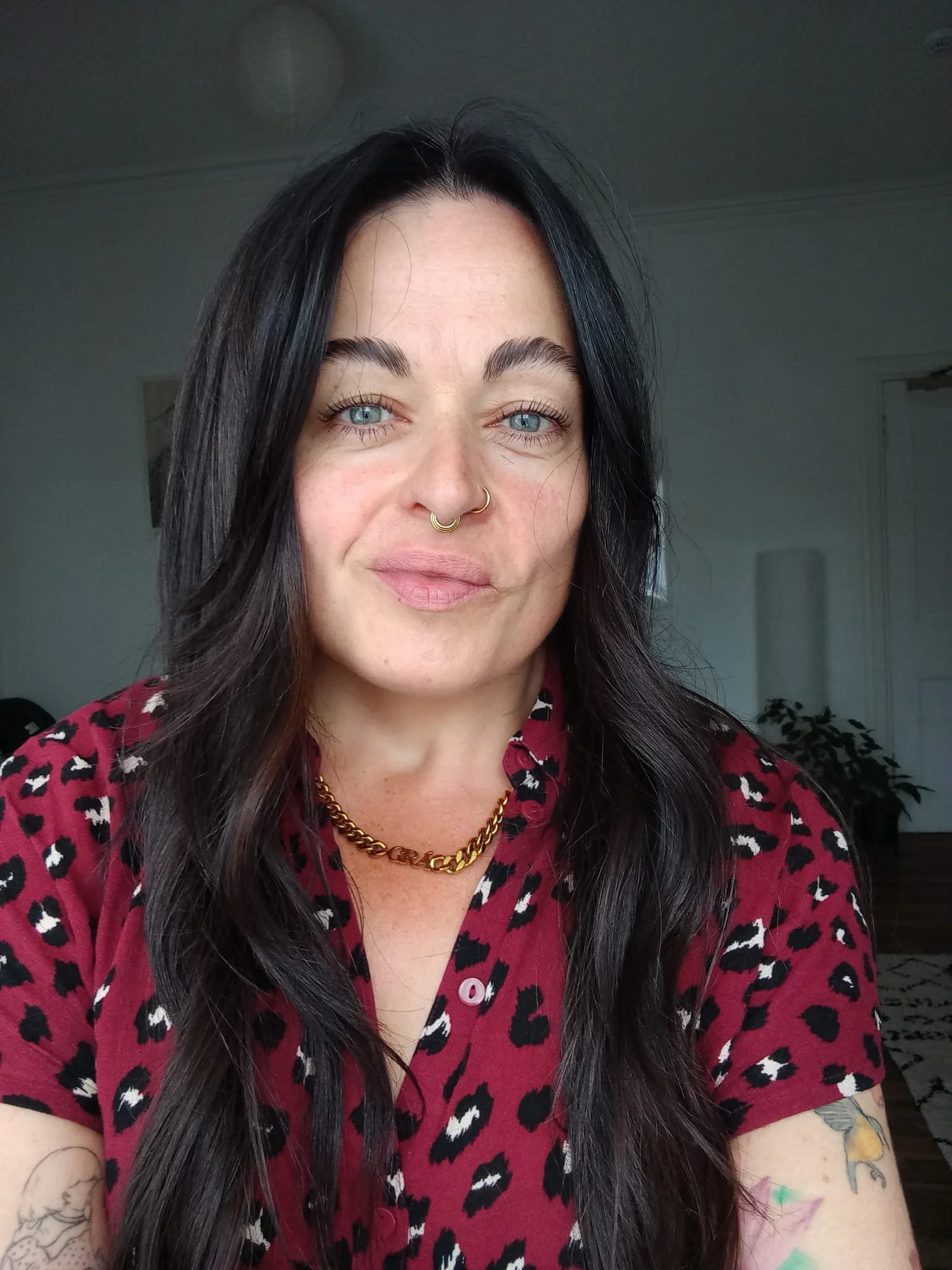 Jules Harringont (she/they) with black hair, blue eyes, and a nose ring taking a selfie indoors. Wearing a red, short-sleeved shirt with a leopard print pattern, a gold chain necklace, and has tattoos on their arms.