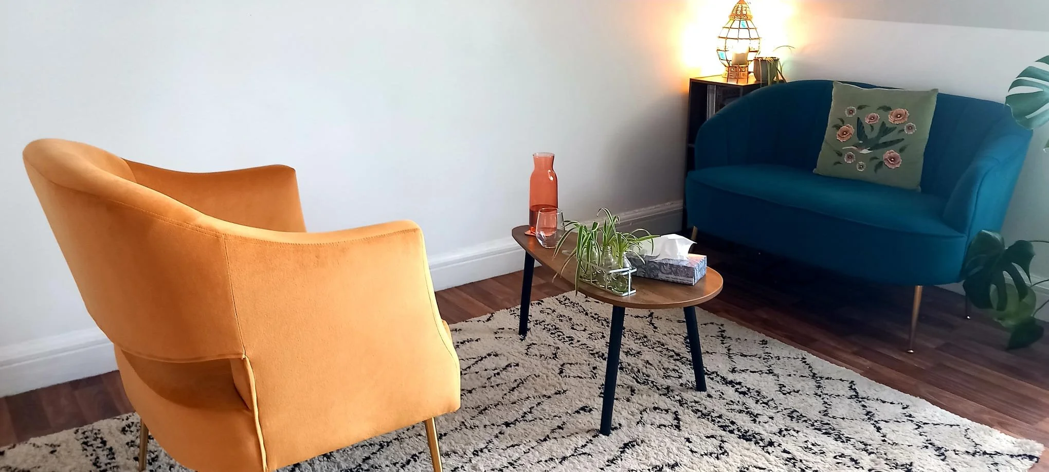 Living room with an orange armchair, a teal loveseat with a floral pillow, a small wooden coffee table with a plant, tissue box, and glass container, a rug with a geometric pattern, steeped hardwood floors, a black bookshelf, and a lamp with a woven lampshade.