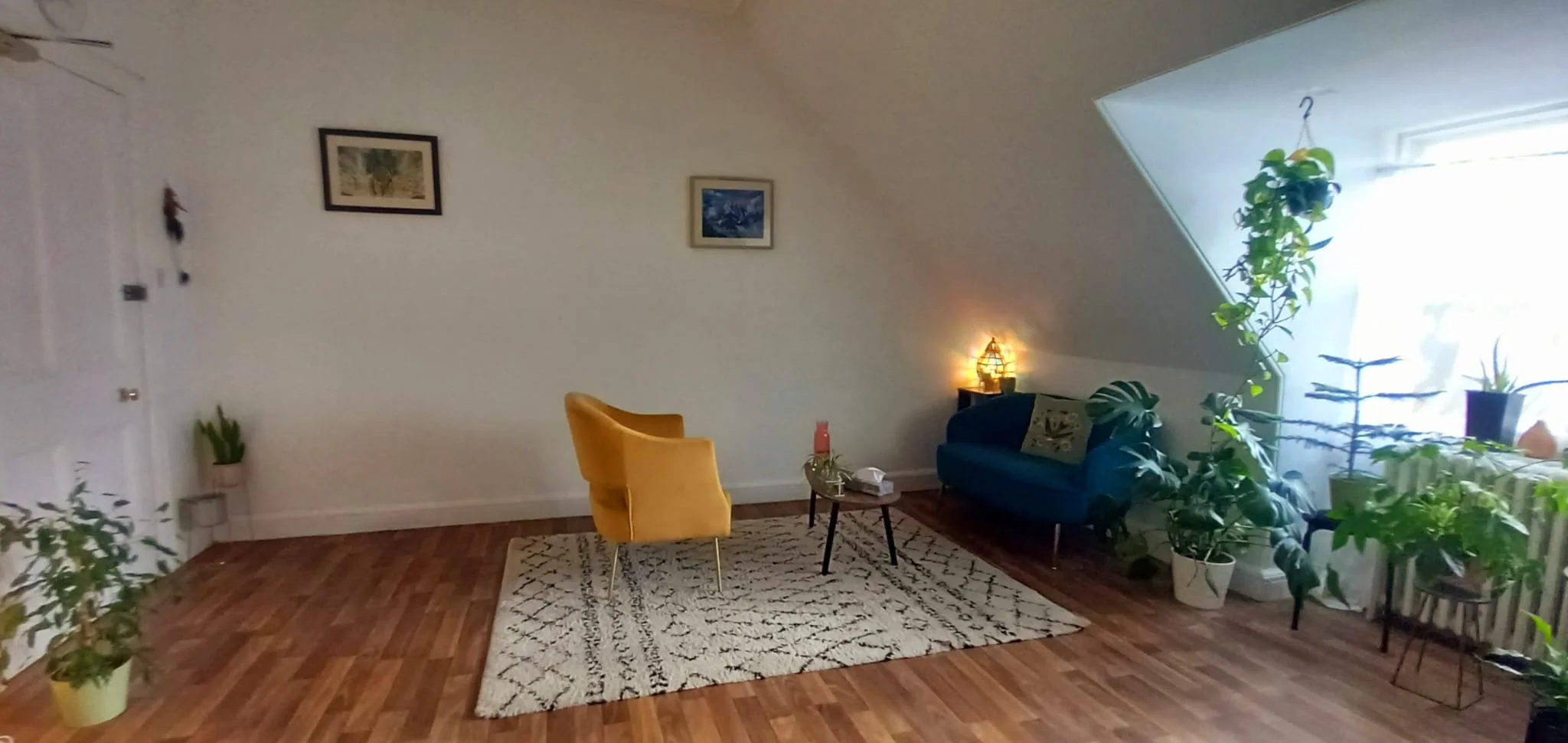 Living room with hardwood floors, a yellow armchair, a blue sofa, plants near a skylight, a patterned rug, and framed pictures on the wall.