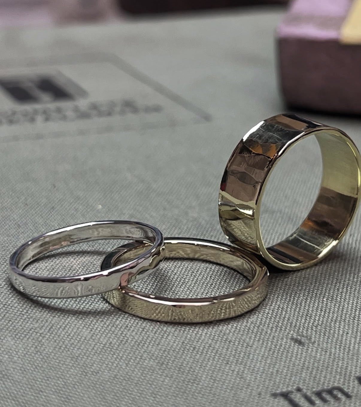 a thing silver ring band placed a quarter of the way over the same sized brass ring band. To the right is a thicker ring band standing on edge. The three rings sit on a sage green book cover that reads as fabric.