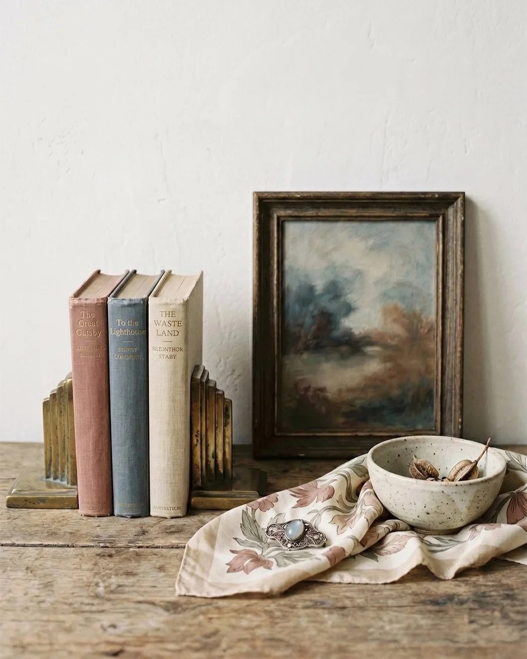 Vintage still life of found objects — brass bookends, cloth-bound antique books, an original landscape painting, a silk scarf, and a pearl brooch — sourced by Second Bloom Style in Chicago.