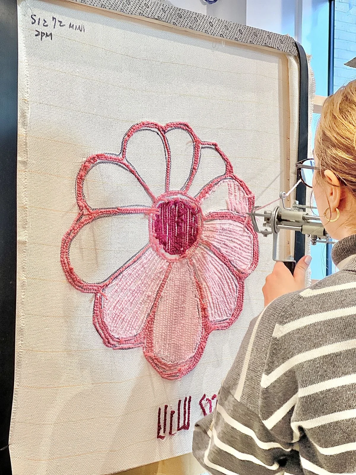 A person using a tufting gun to work on a large, pink embroidered flower fabric art hanging on a wall.