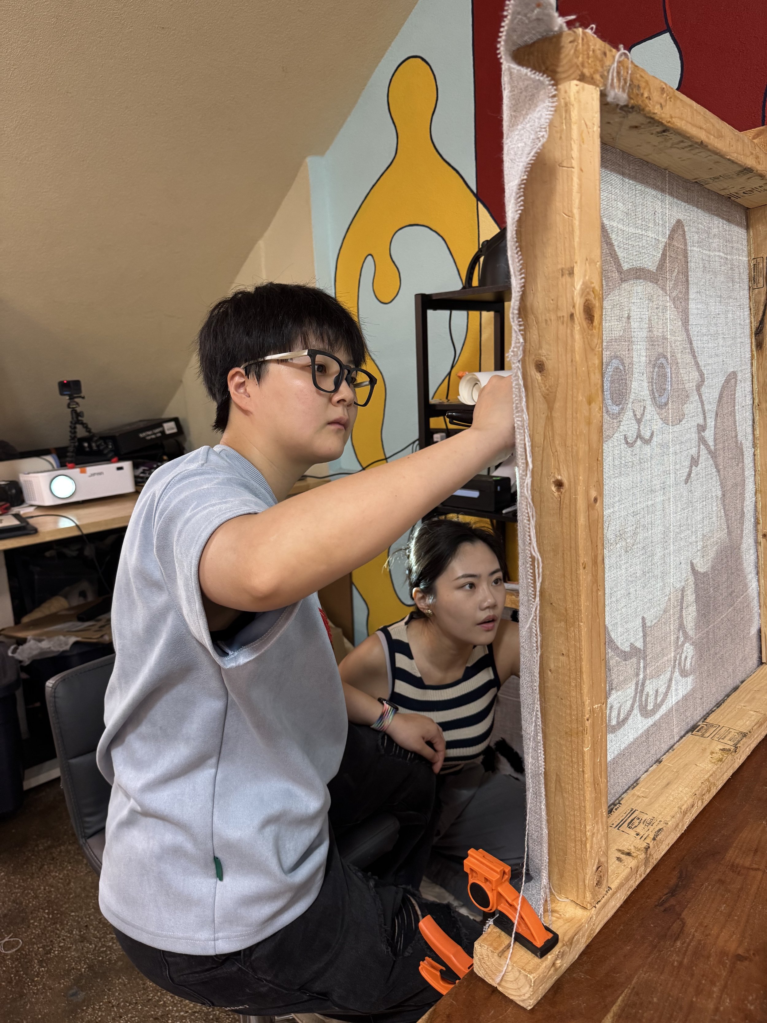 Two people working on a tu project inside a room with colorful wall art, one person wearing glasses and a gray shirt, the other sitting next to them watching. The workspace includes a wooden frame with fabric, tools, and a small orange plastic clamp.