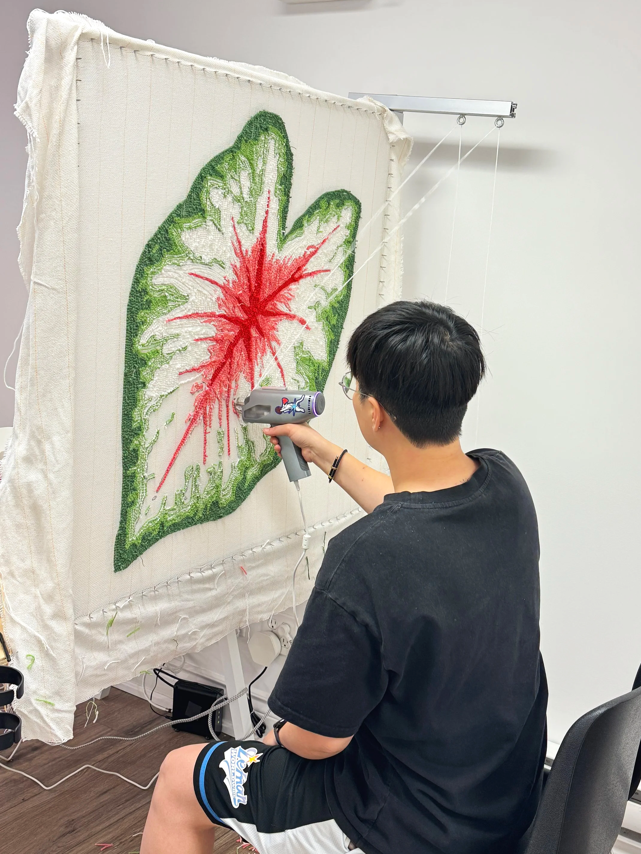 A person sitting in a black chair, working on a large tufting piece with a colorful leaf pattern at Dotty Tuft Studio