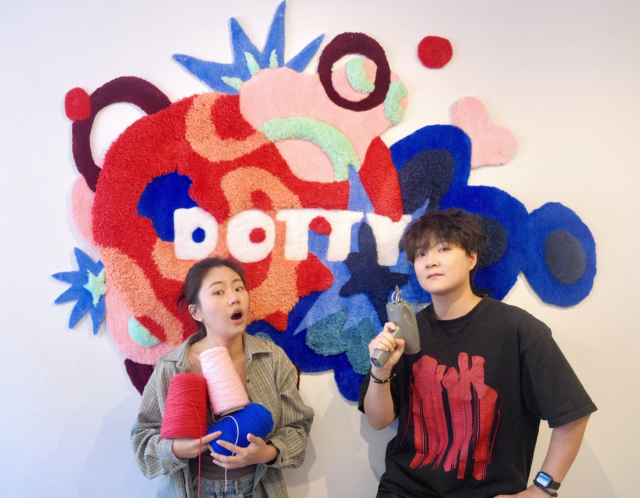 Two people standing in front of a colorful tufting fabric wall art with the word 'Dotty'.