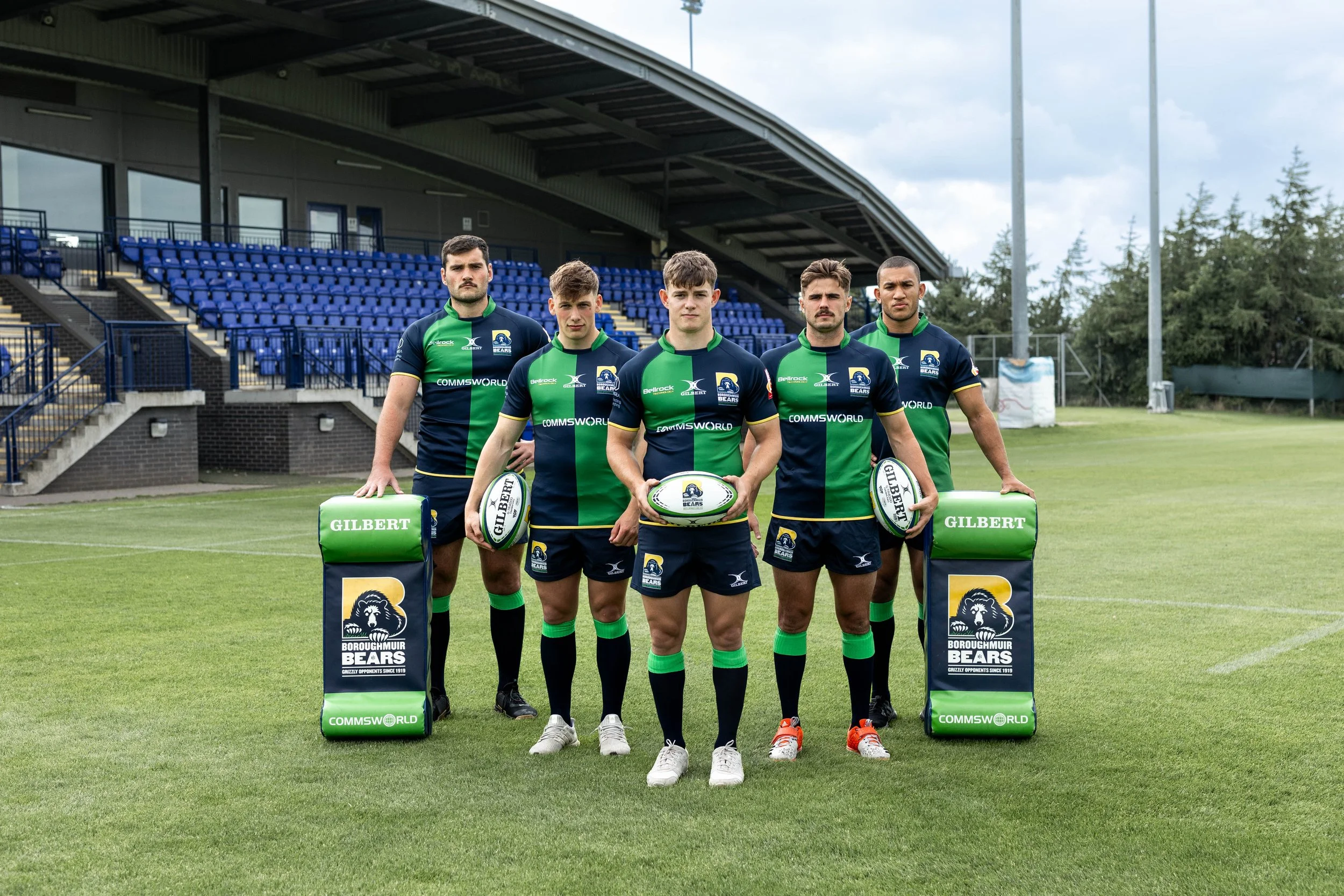 Five rugby players stand on a field at their rugby club in the uae in their Gilbert rugby kit, holding Gilbert rugby balls and Gilbert rugby equipment, with a stadium seating and goalposts in the background.