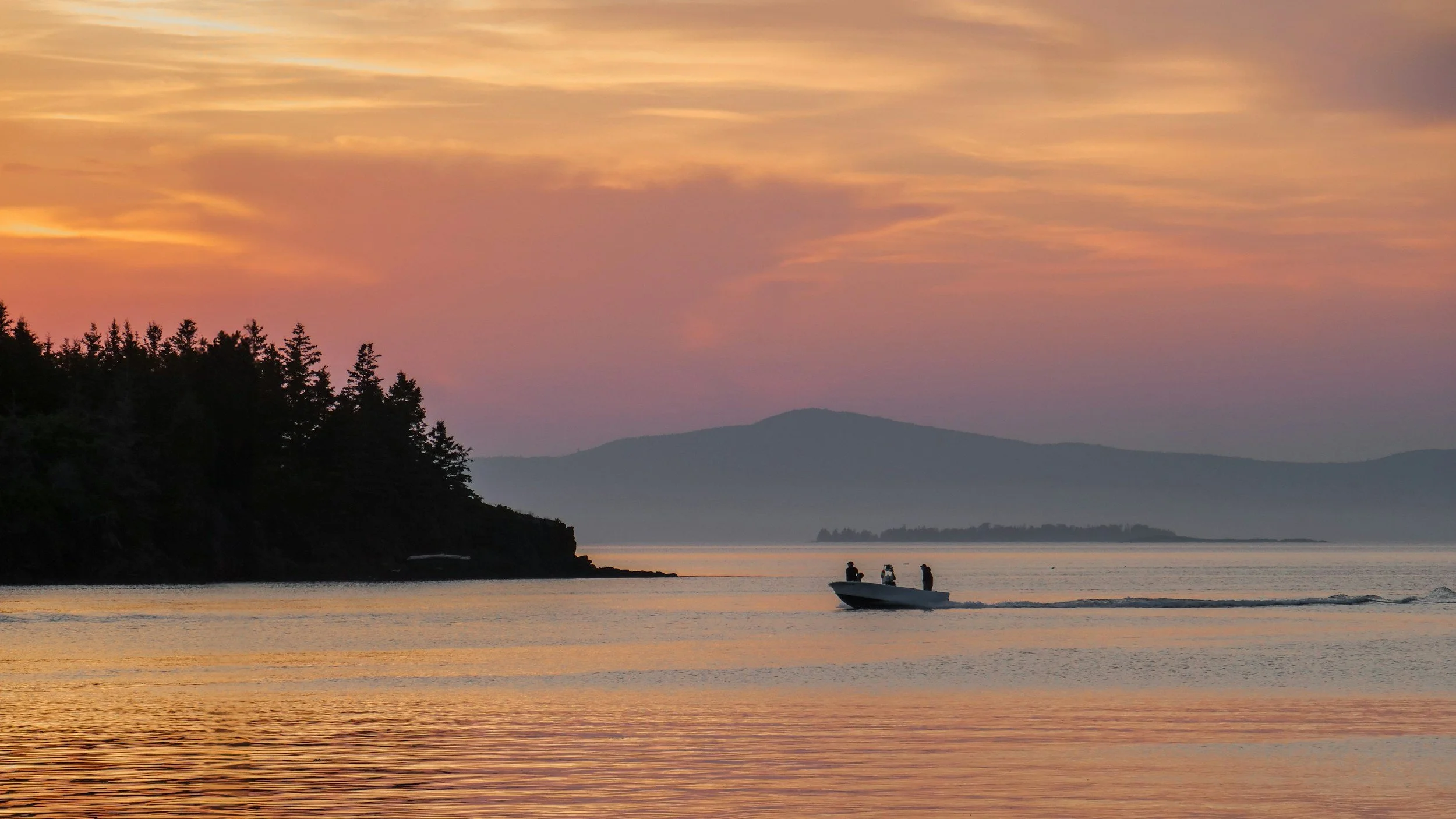 A boat with three people on it is moving across a calm body of water during a sunset, with a silhouette of trees on the left and mountains in the background, under a colorful sky with orange, pink, and purple hues.