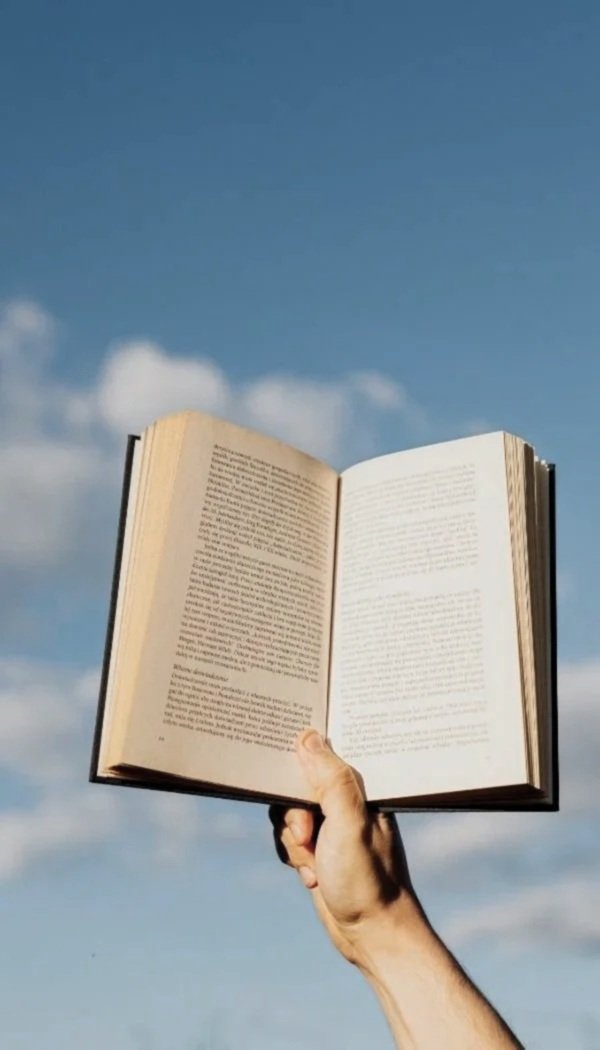 Een arm houdt een open boek omhoog tegen een blauwe hemel met enkele wolken.