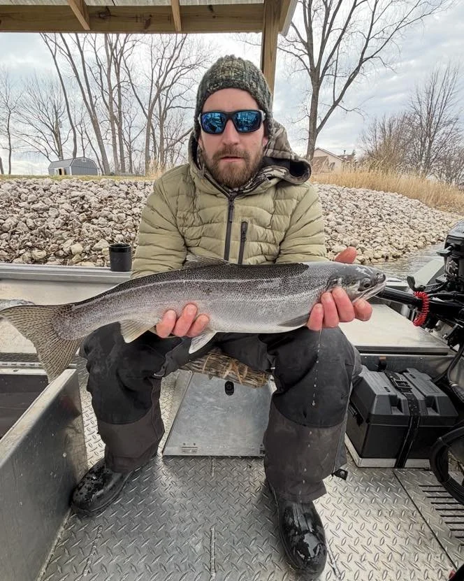 Dakota the owner of fancy fish charters is wearing a camouflage beanie, a tan quilted jacket, and waterproof pants is sitting in a boat holding a large fish with both hands. The boat is on water, with trees and a rocky shoreline in the background.