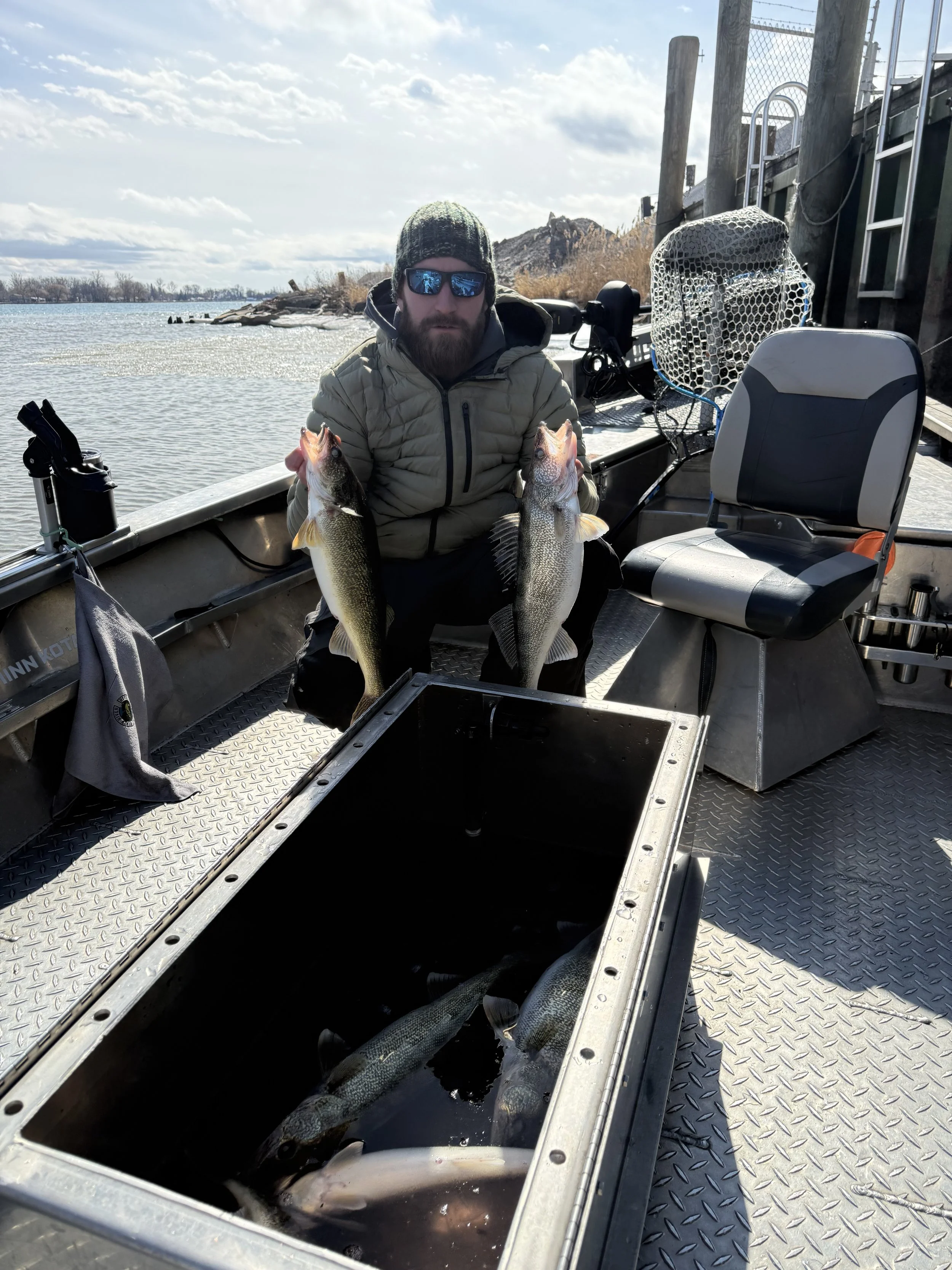 Man in a beige jacket, black cap, and reflective sunglasses holding two large fish on a boat near a body of water with a rocky shoreline in the background.