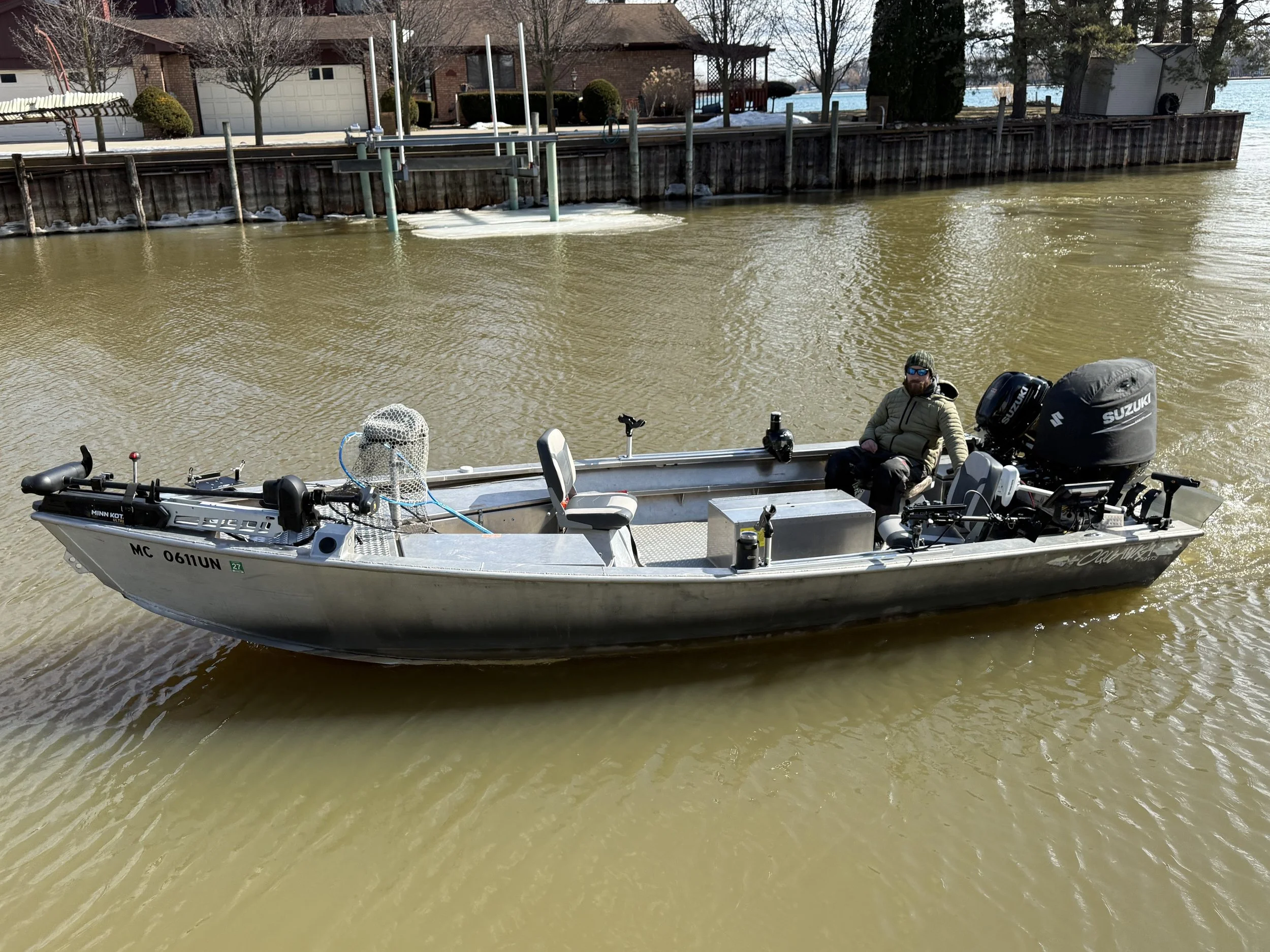 A small motorized boat with multiple items on board, including a person wearing sunglasses and a hat, is floating on a waterway near a wooden and metal dock with houses in the background. The boat has Suzuki outboard motors and various equipment.