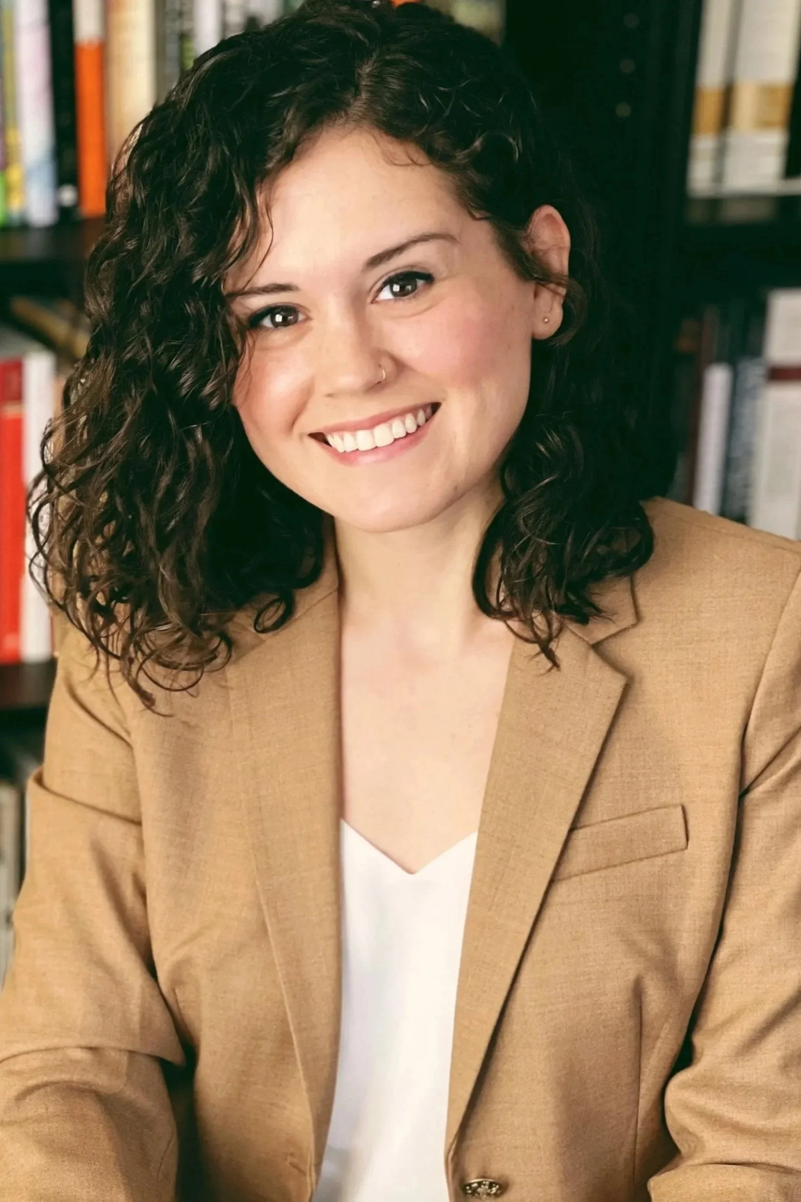 Close-up of a Latina woman with curly dark hair and dark brown eyes, wearing a tan blazer over a white top, smiling, with bookshelves in the background.