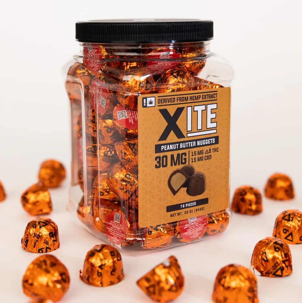 A clear plastic jar with a black lid, filled with orange foil-wrapped peanut butter nuggets labeled as XITE, derived from hemp extract, containing 30 mg per piece with 7 pieces in total. The jar is surrounded by a few more unwrapped or partially unwrapped nuggets