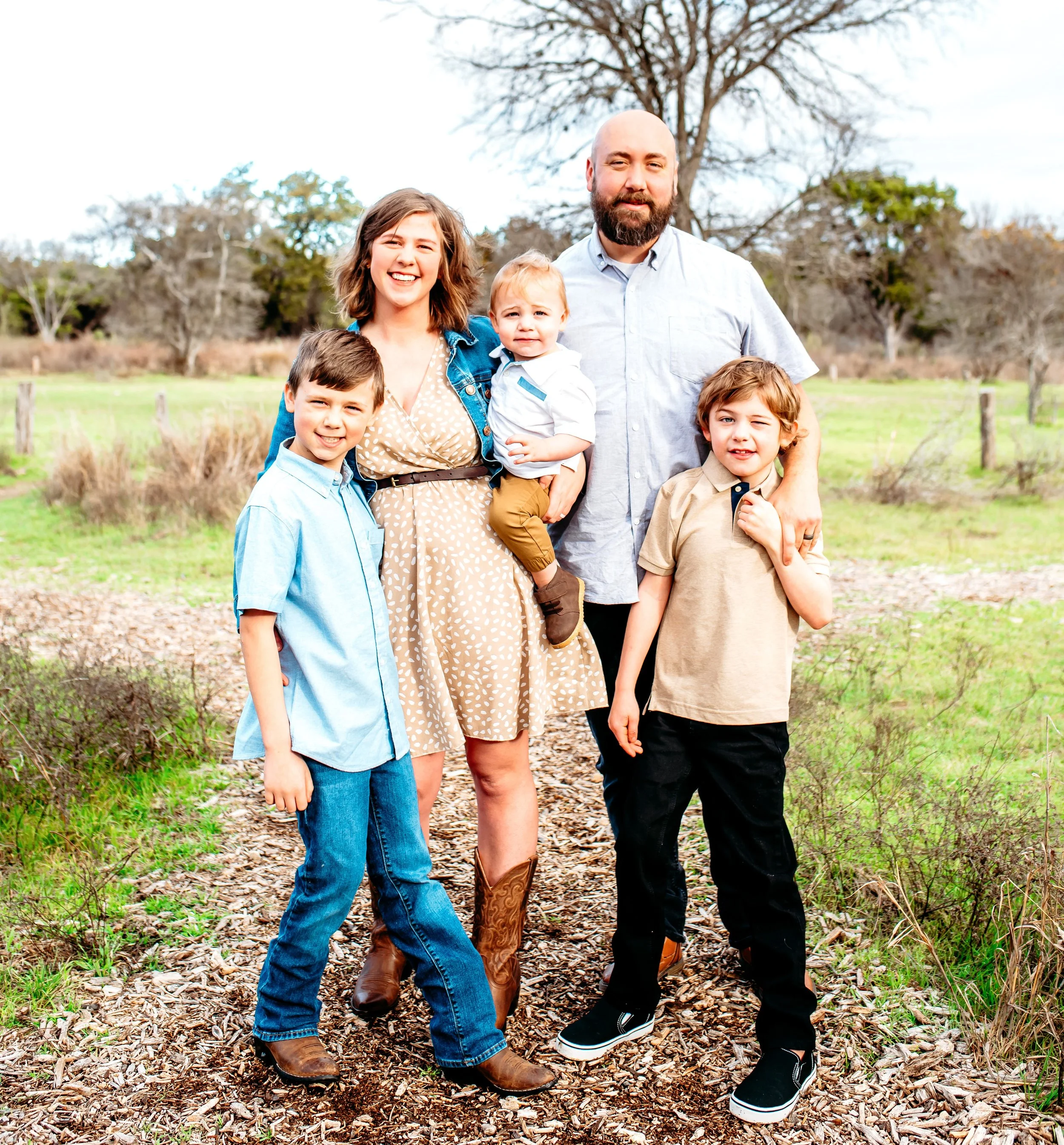 A group of six people, including three children and three adults, standing on a trail in a park or wooded area with leafless trees in the background. They are smiling and dressed casually for outdoor weather.
