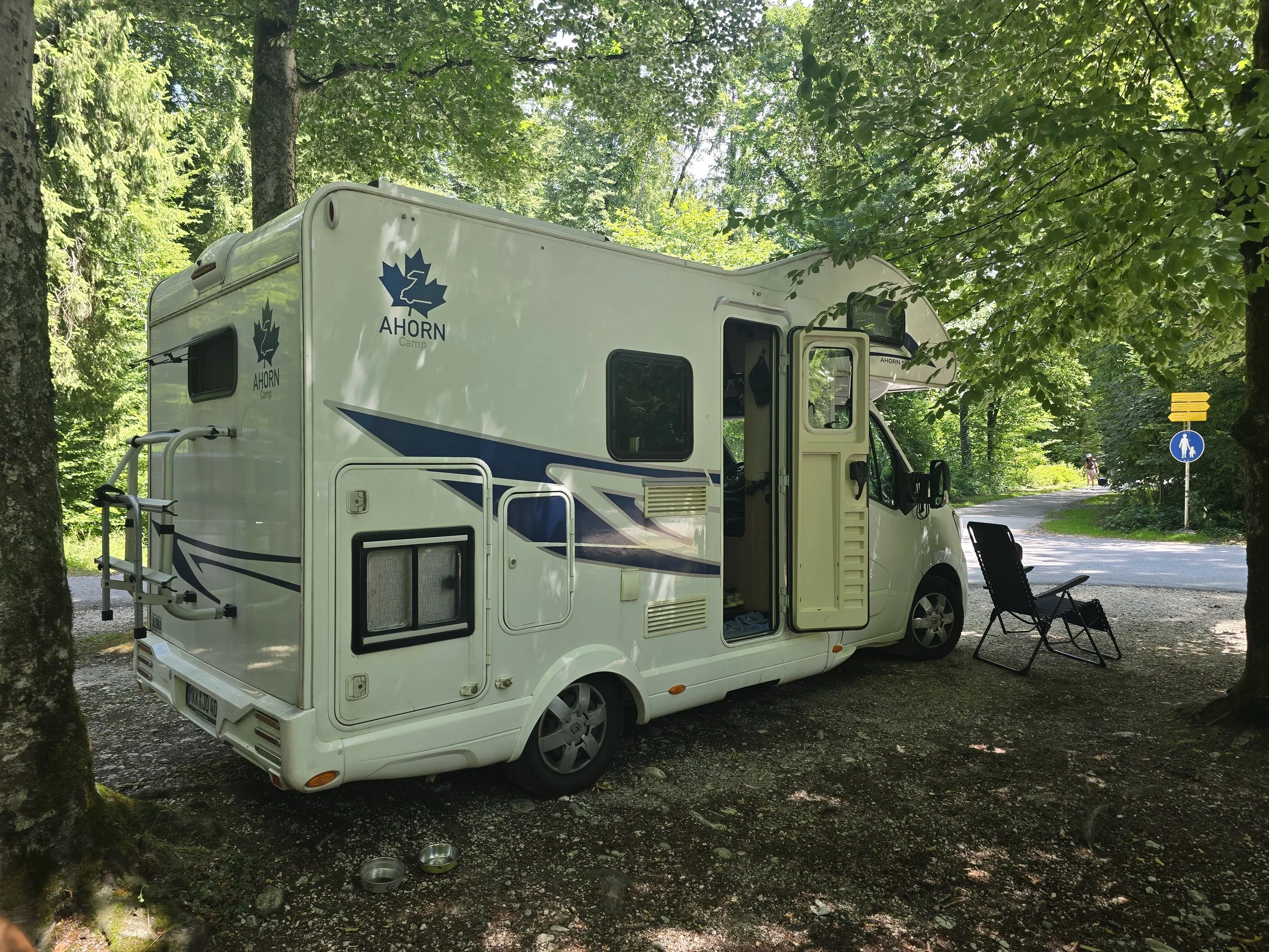Alt-Text: Weißes Wohnmobil auf einem schattigen Stellplatz unter Bäumen in Bad Reichenhall, daneben steht ein Campingstuhl.