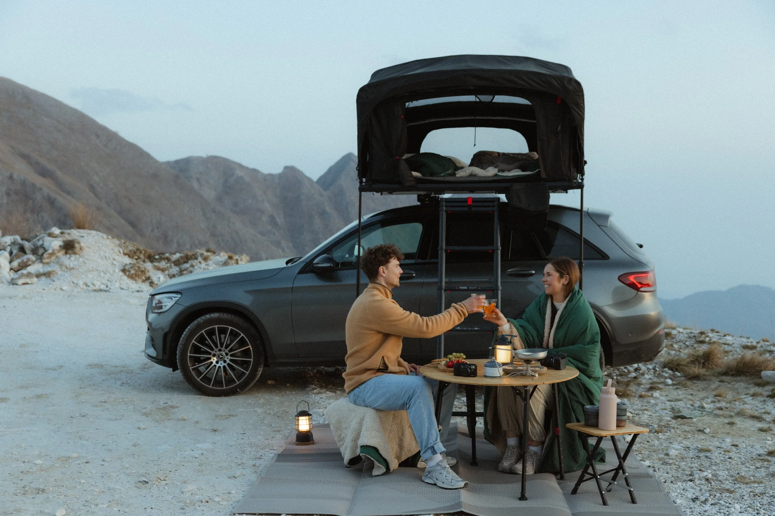 Paar sitzt vor einem Auto mit aufgebautem Dachzelt an einem Campingtisch in den Bergen.