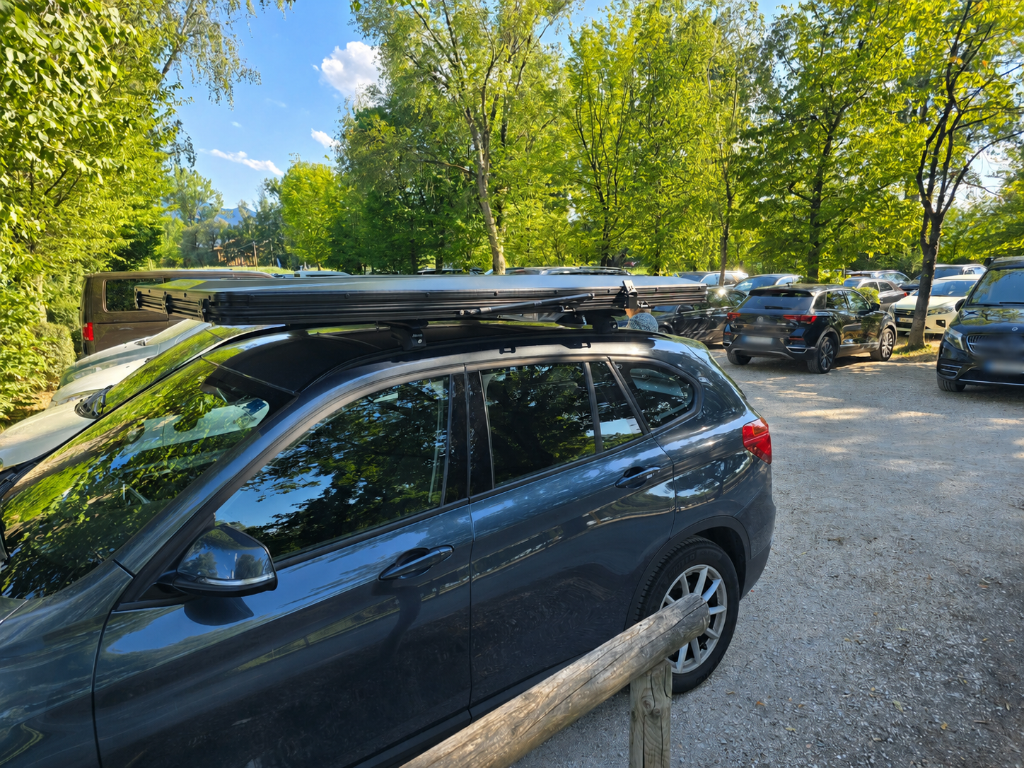 Zusammengeklapptes Dachzelt auf einem dunklen Auto mit Querträgern auf einem Parkplatz im Grünen.
