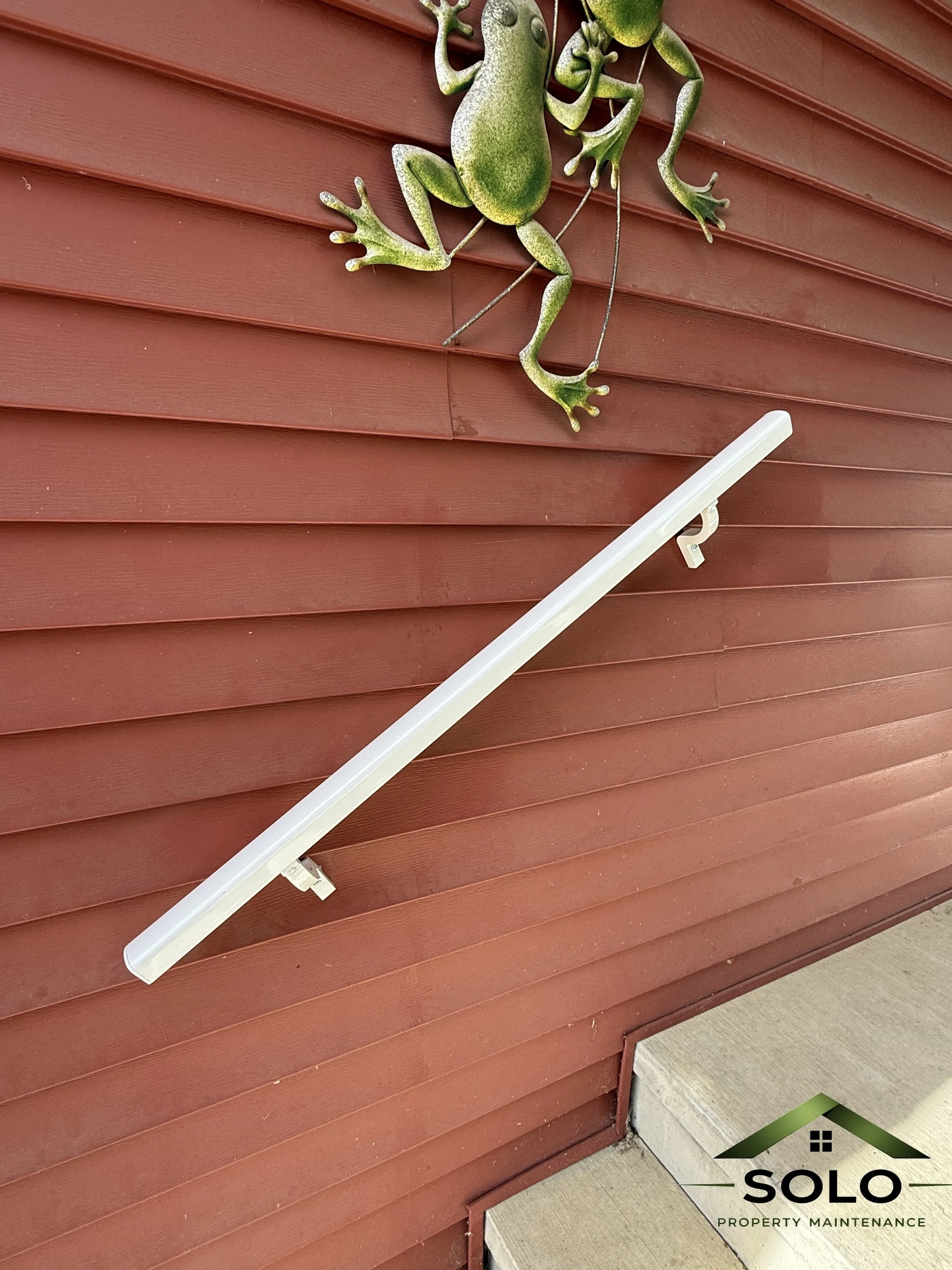 A decorative green gecko wall ornament on a red wooden wall beneath a white siding. A white solar-powered outdoor light fixture is attached below the gecko. The SOLA Property Maintenance logo is visible in the bottom right corner.