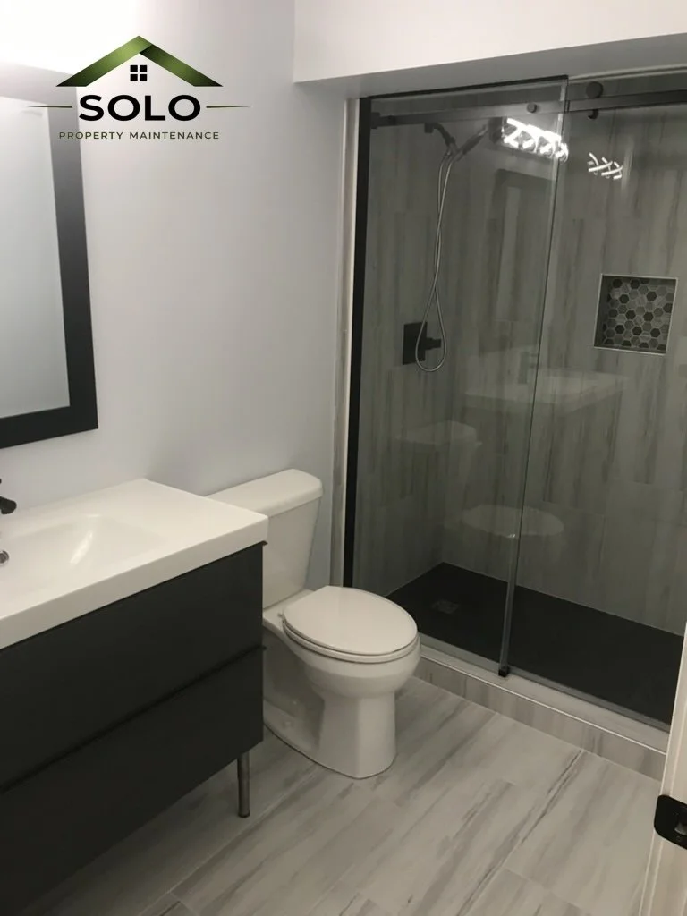 Modern bathroom with a white toilet, black vanity with white sink, and a glass-enclosed shower with wood-look tiles and a built-in niche.