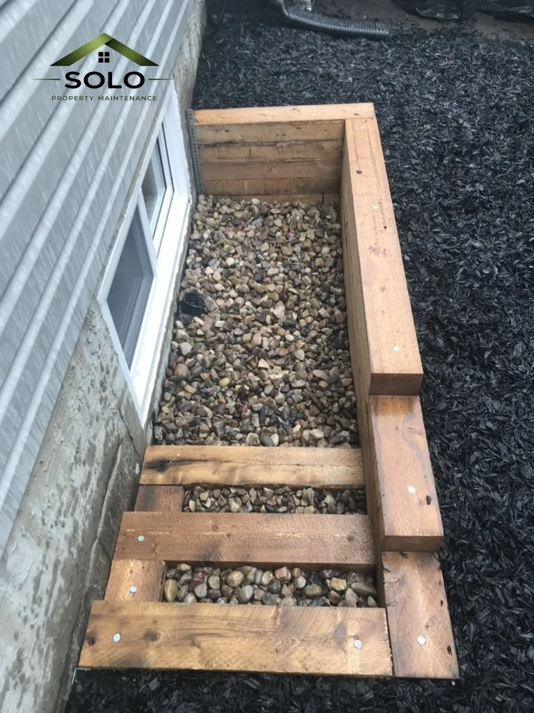A wooden frame structure under construction next to a house with a window, filled with small rocks, and surrounded by black mulch.