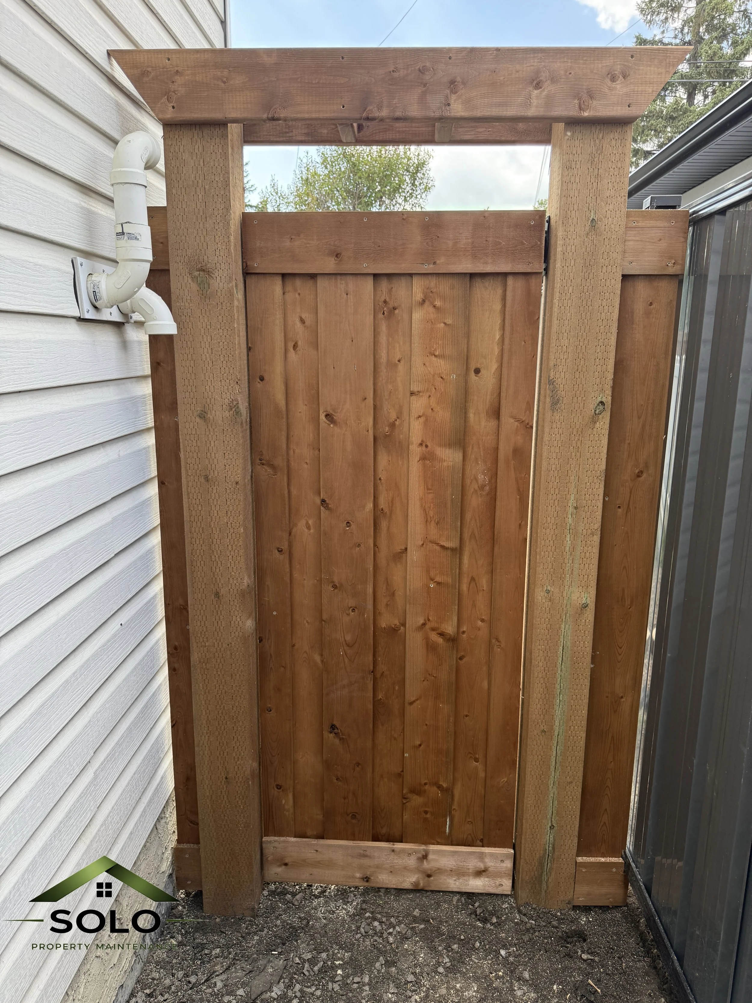 A newly built wooden privacy fence with gate, situated between a house with white siding and a glass enclosure, on a gravel ground background under a blue sky with clouds.