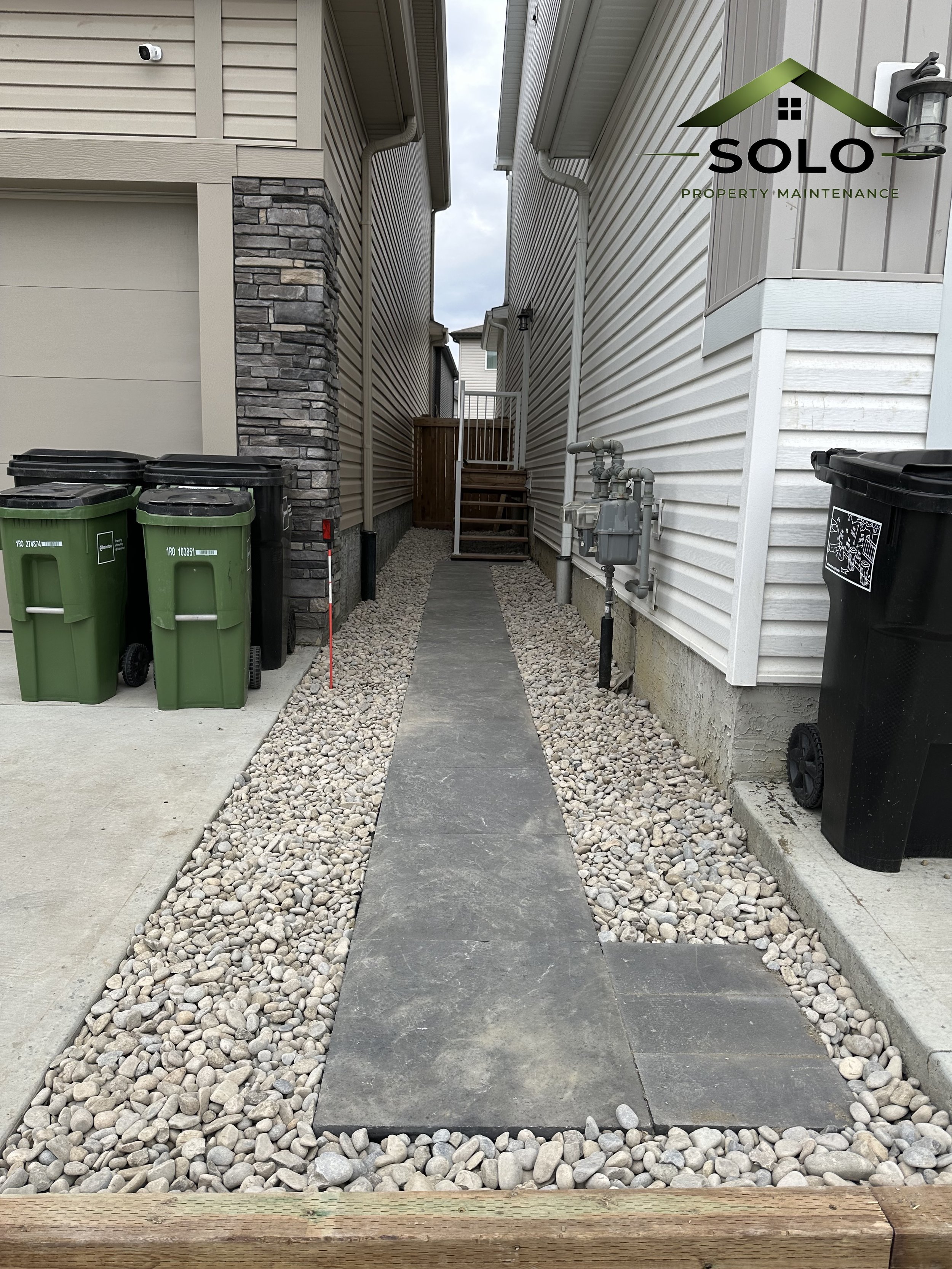 A narrow outdoor passageway between two houses with gravel on the sides and a concrete and stone path leading to a set of stairs with a wooden gate at the top.