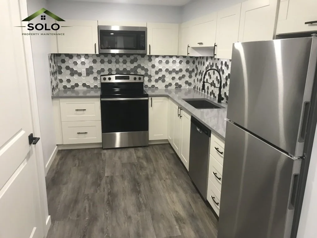 Modern kitchen with white cabinets, gray hexagonal tile backsplash, black and stainless steel appliances, gray patterned countertop, and wood-look flooring.