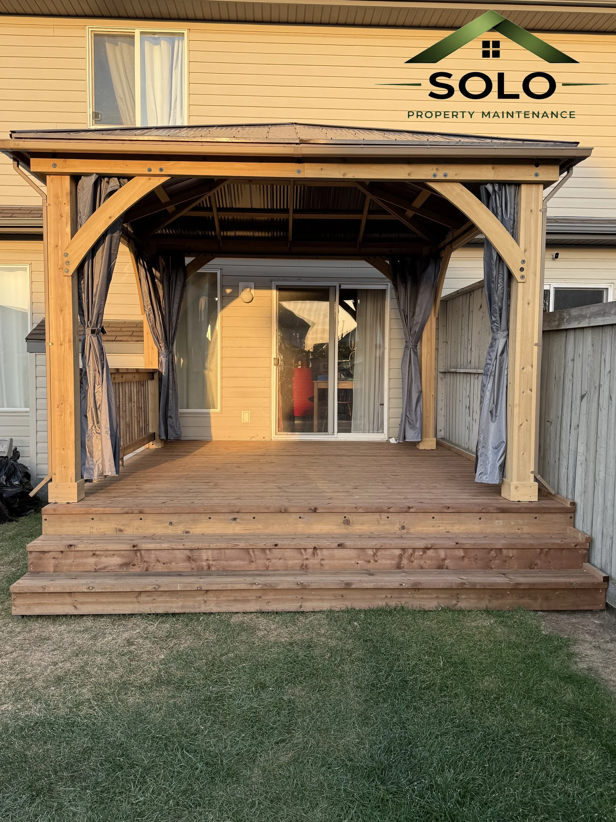 A wooden deck attached to the back of a house, with a pergola framework and curtains, and a sliding glass door leading inside.