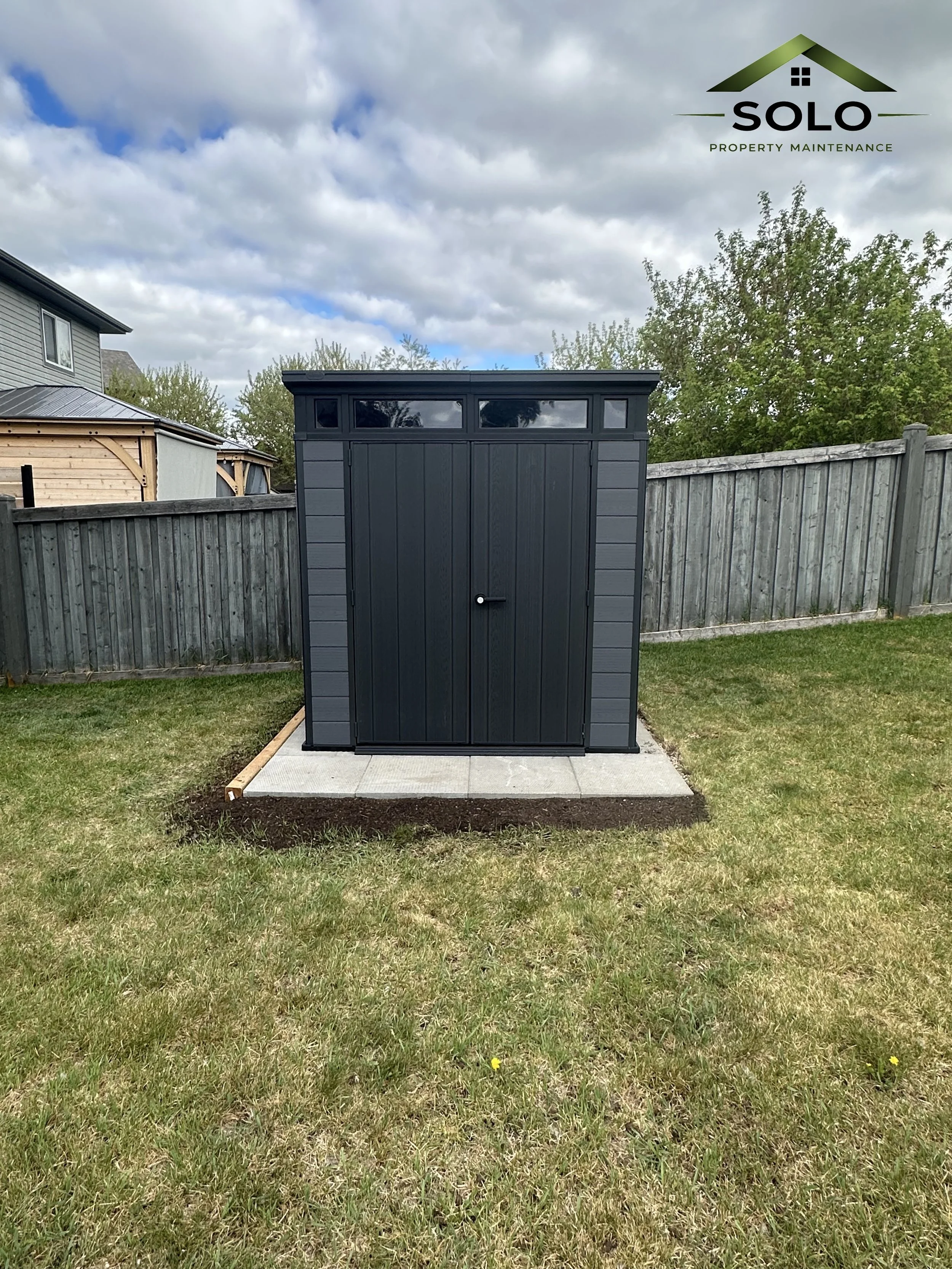 A small, black shed with a sloped roof and a door, situated on a concrete pad in a grassy backyard enclosed by a wooden fence. There are trees and neighboring houses in the background, with a cloudy sky overhead. The image has a logo for Solo Propert