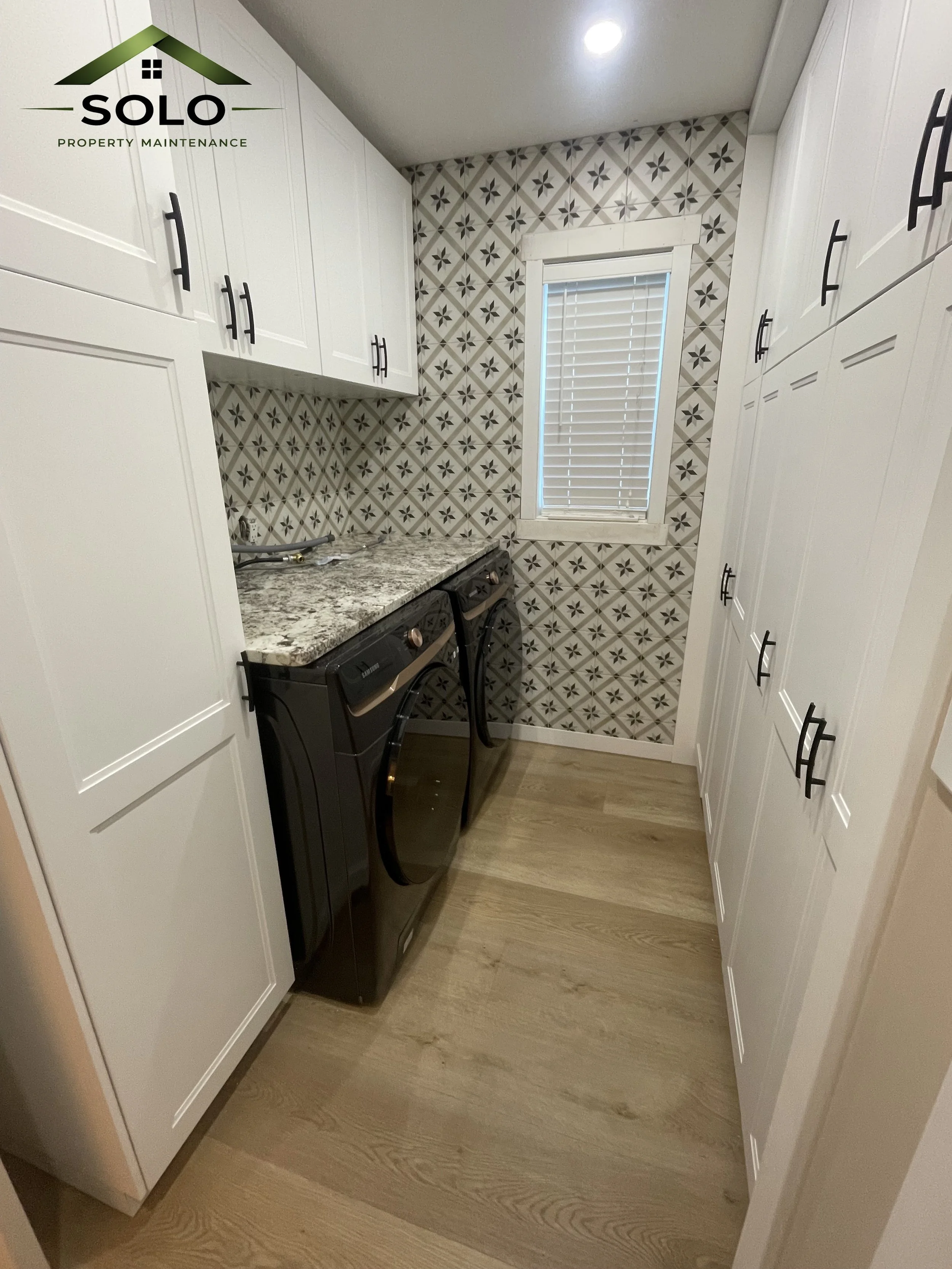 A small laundry room with white cabinets, a granite countertop, and patterned tile walls. A window with blinds is centered on the back wall, and a black washing machine is underneath the counter. There is wood flooring and a ceiling light.