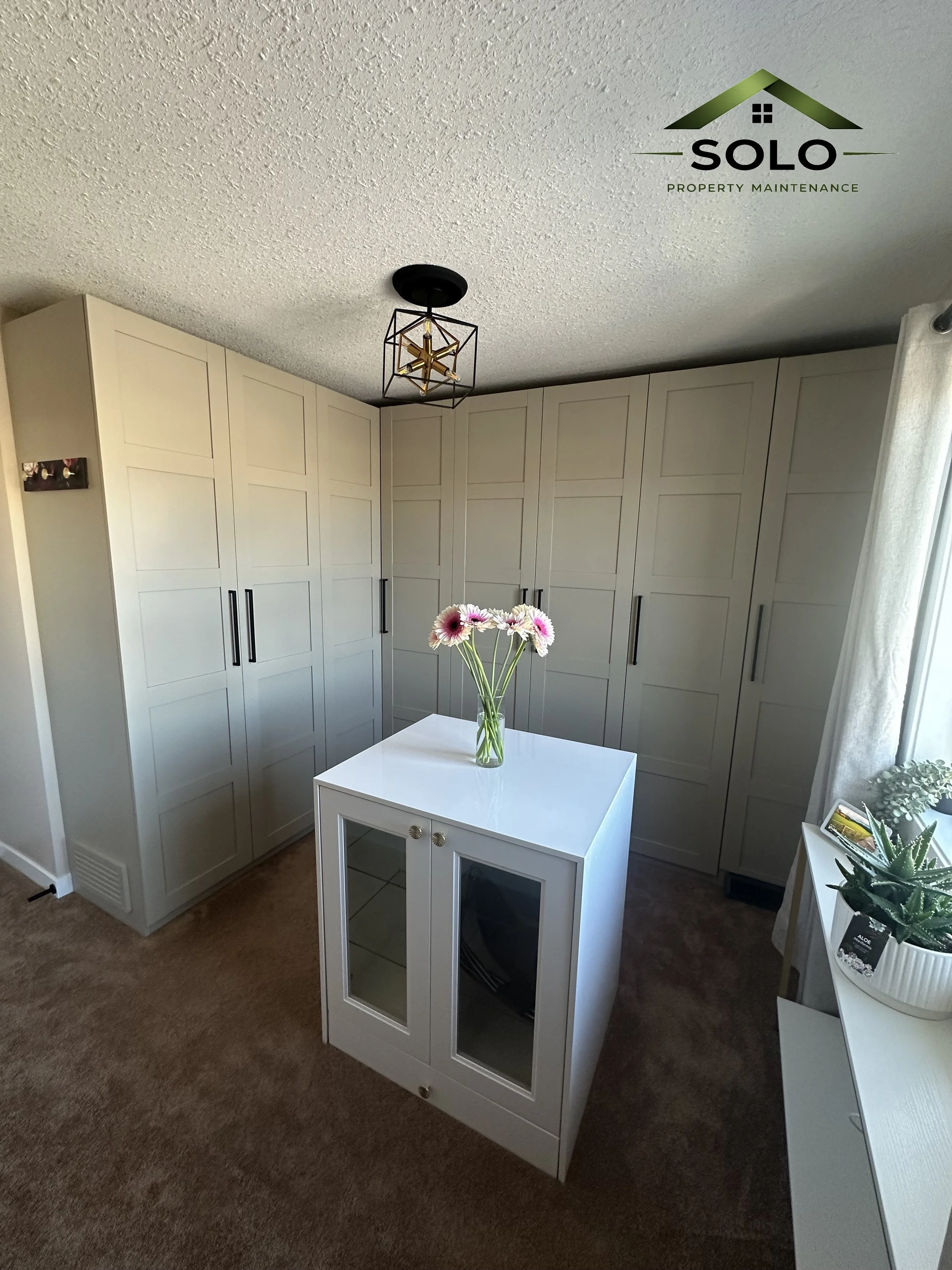 A corner of a room with white cabinets with black handles, a small white table with a glass door oven or storage space, a vase of pink and white flowers, a potted plant on a white stand, and a window with white curtains. There is a modern ceiling lig