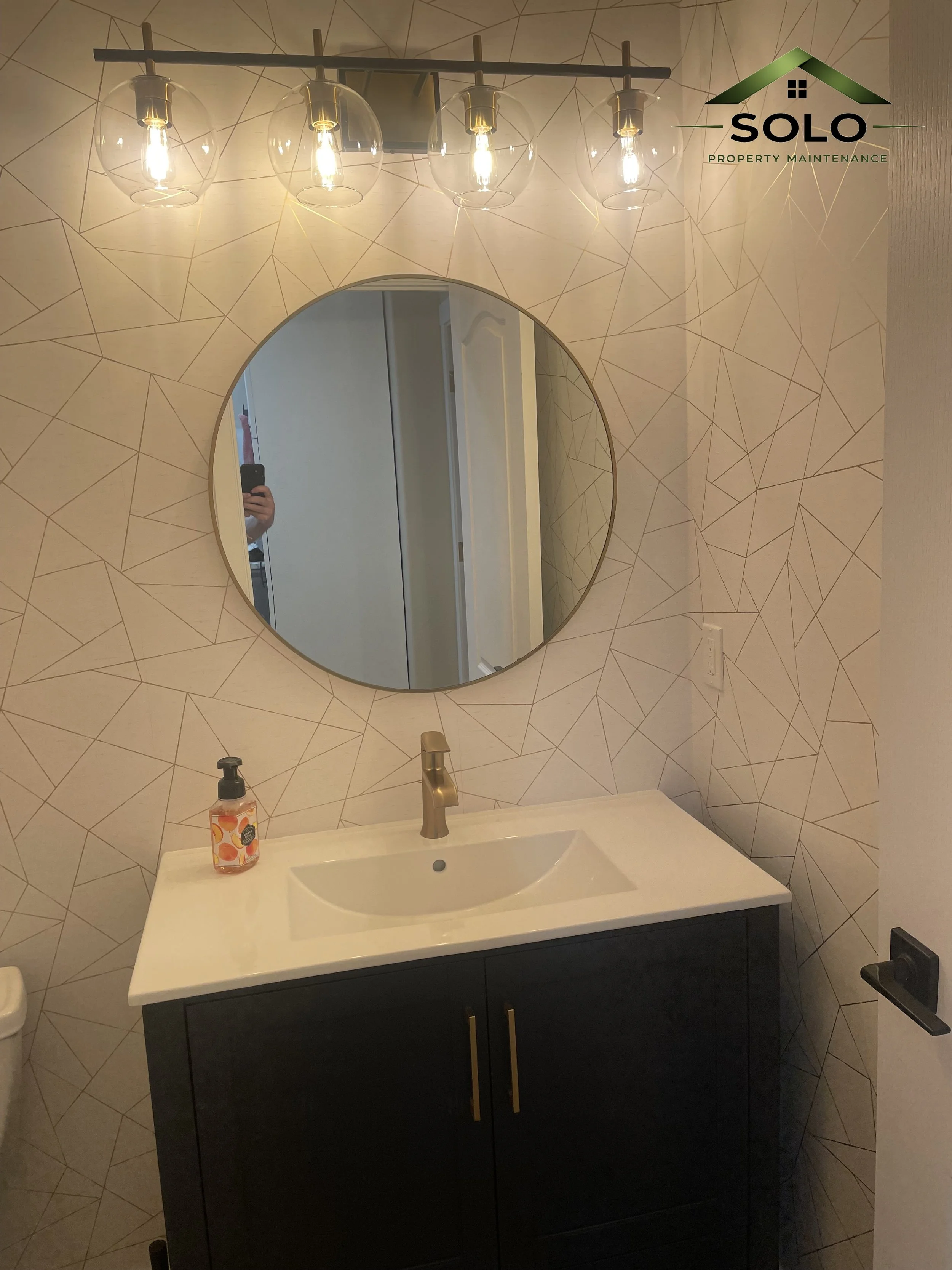 A bathroom sink with a black vanity and gold handles, a white countertop, and a gold faucet. A bottle of soap with orange and yellow fruit design is on the counter. There is a round mirror above the sink and a modern light fixture with four exposed b