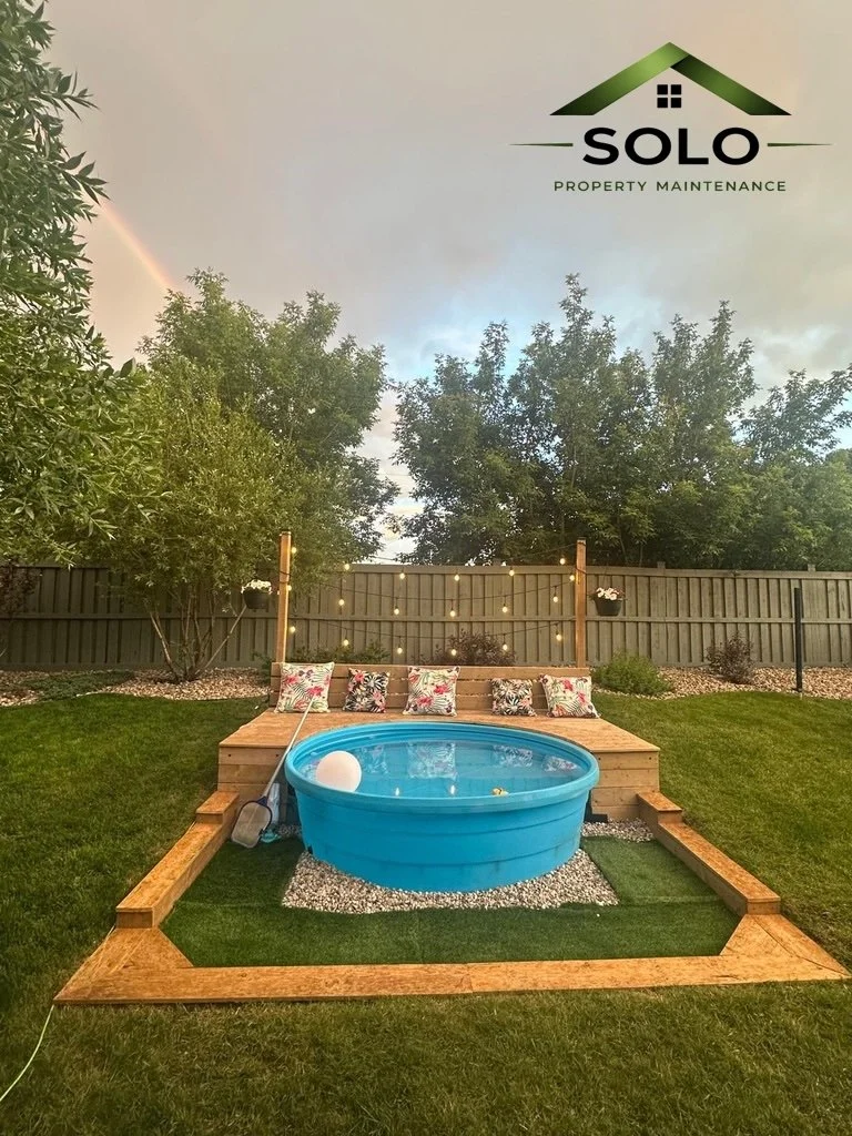 A backyard scene featuring a small blue plastic pool on a gravel base, surrounded by a wooden deck with pillows, a string of hanging lights, and a grassy area with trees and a fence in the background.