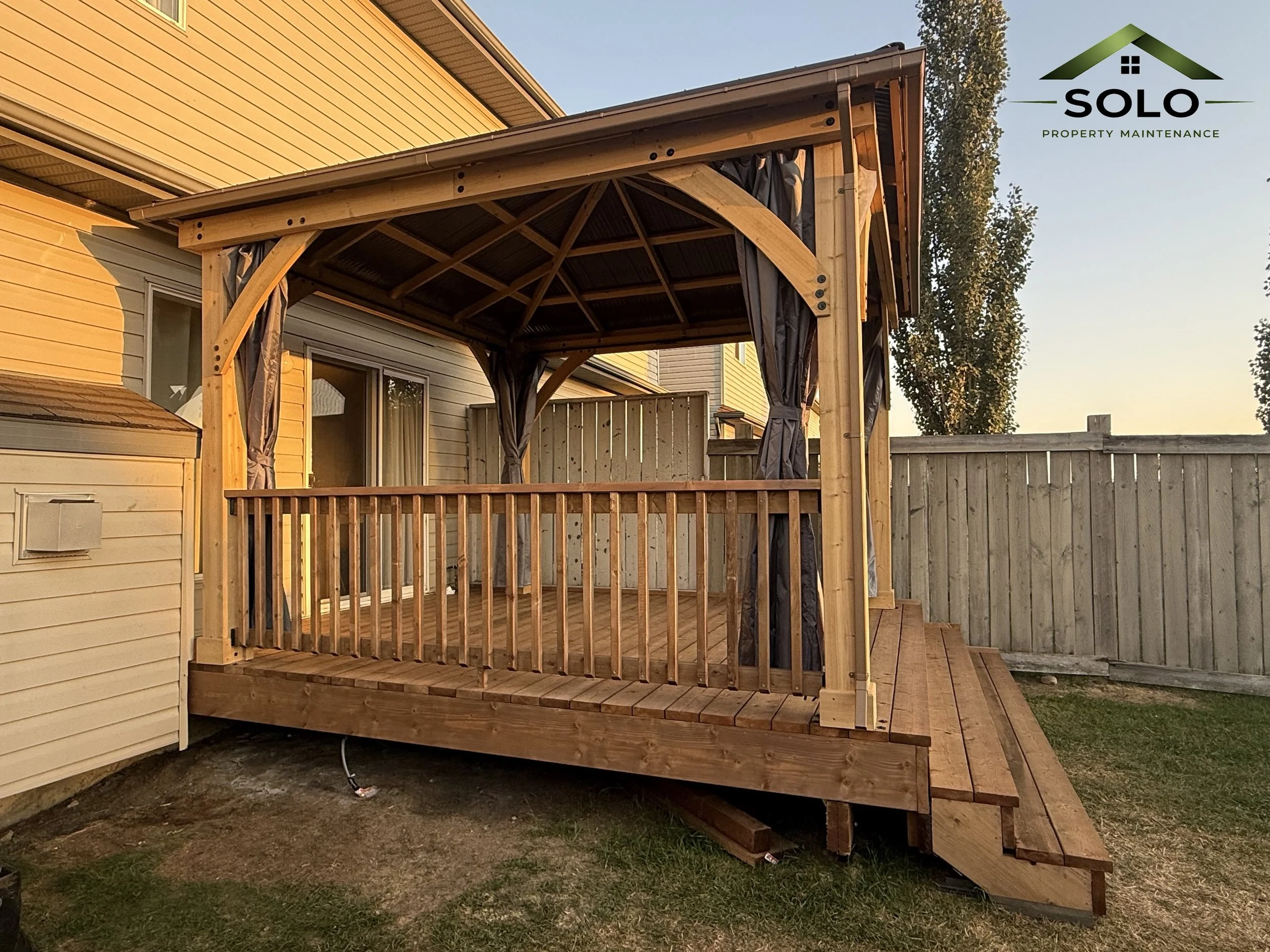 A wooden deck with a privacy fence and curtains, attached to a yellow house with vertical siding, in a backyard with a tree and a clear sky.