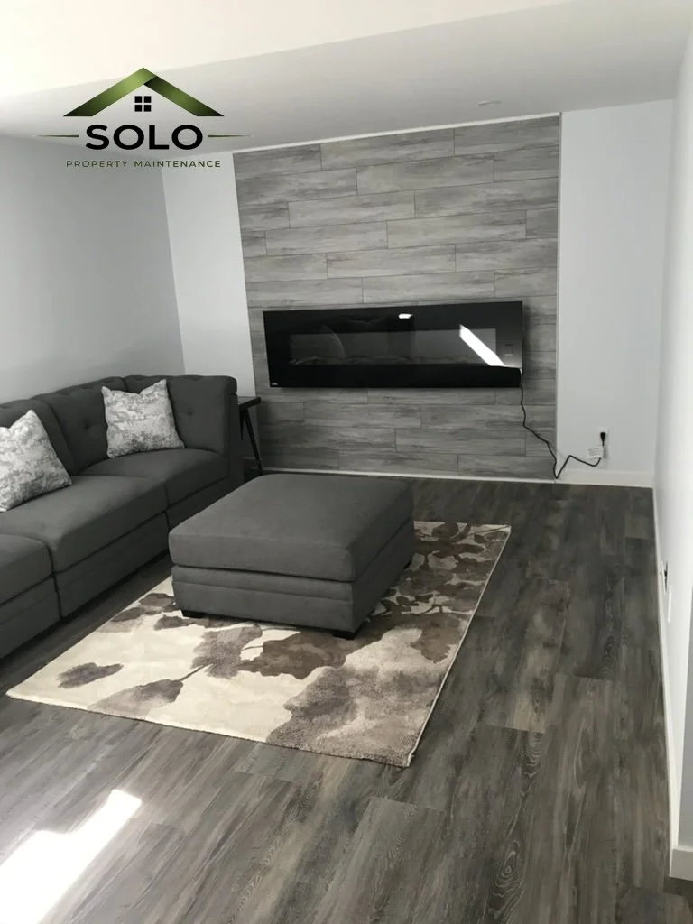 Modern living room with gray sofa, matching ottoman, area rug, wall-mounted fireplace, wood-look flooring, and a decorative wall panel.