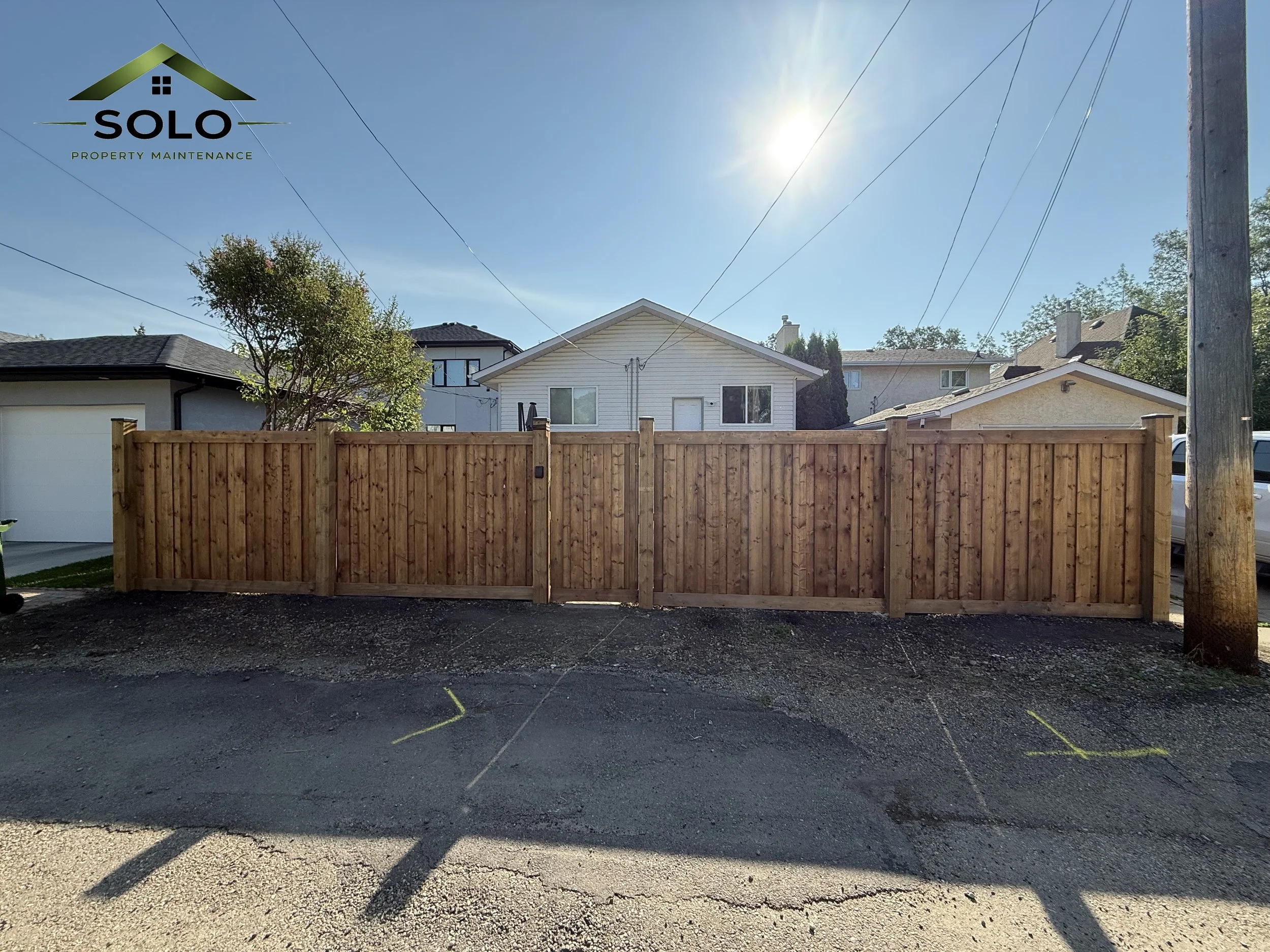 A residential backyard with a wooden fence, a tree, neighboring houses, utility poles, and a clear blue sky with the sun shining brightly.