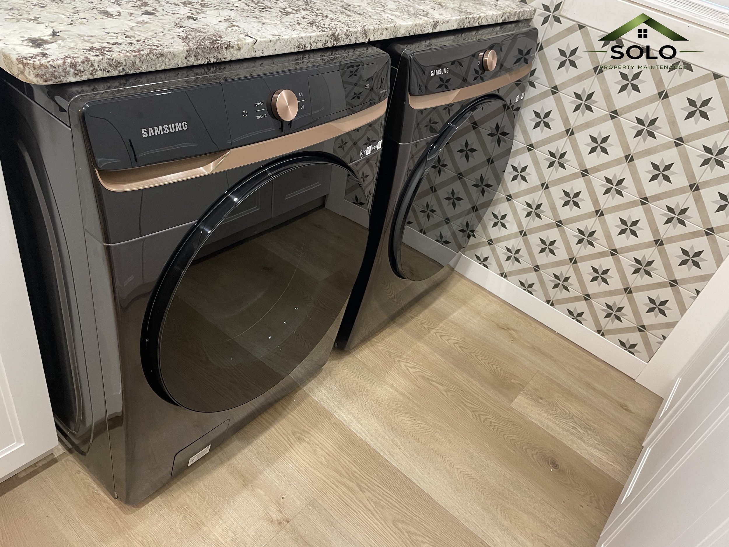 A pair of dark gray Samsung washing machines with beige accents, placed side by side in a laundry room with patterned tile flooring.