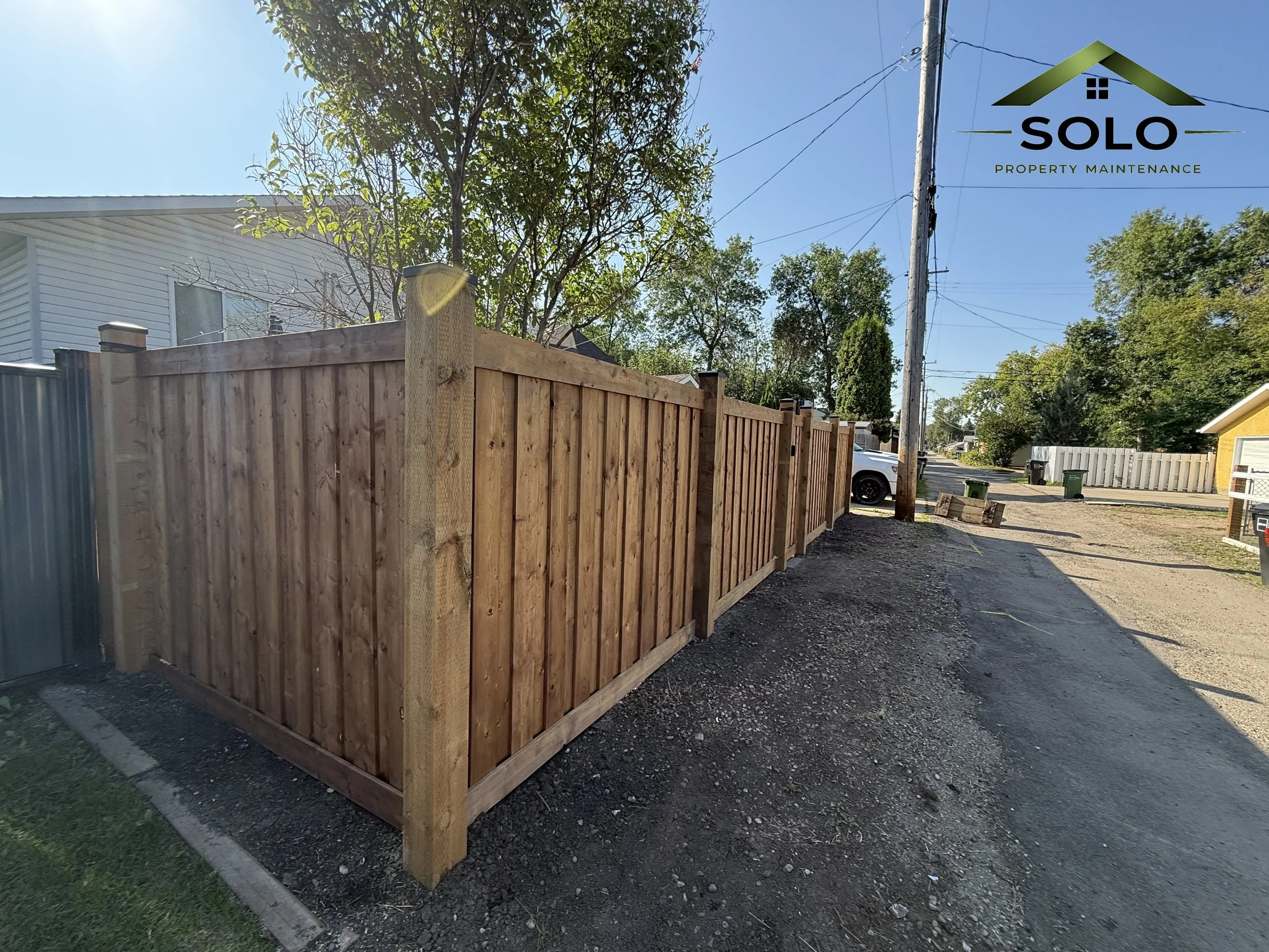New wooden privacy fence along a sidewalk with trees and house in the background on a sunny day.